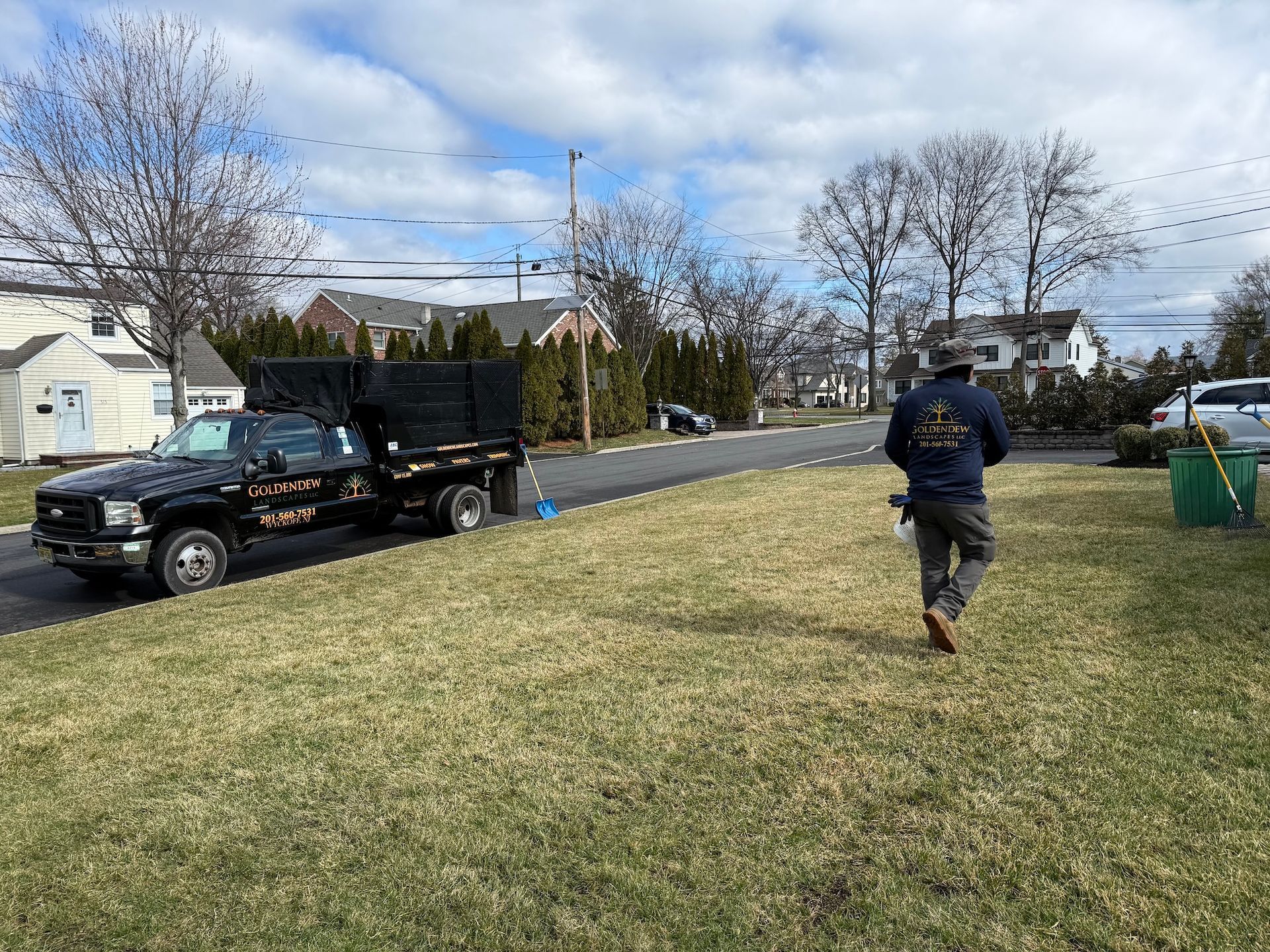 A worker in a dark uniform walks across a lawn near a black dump truck parked on a suburban street.