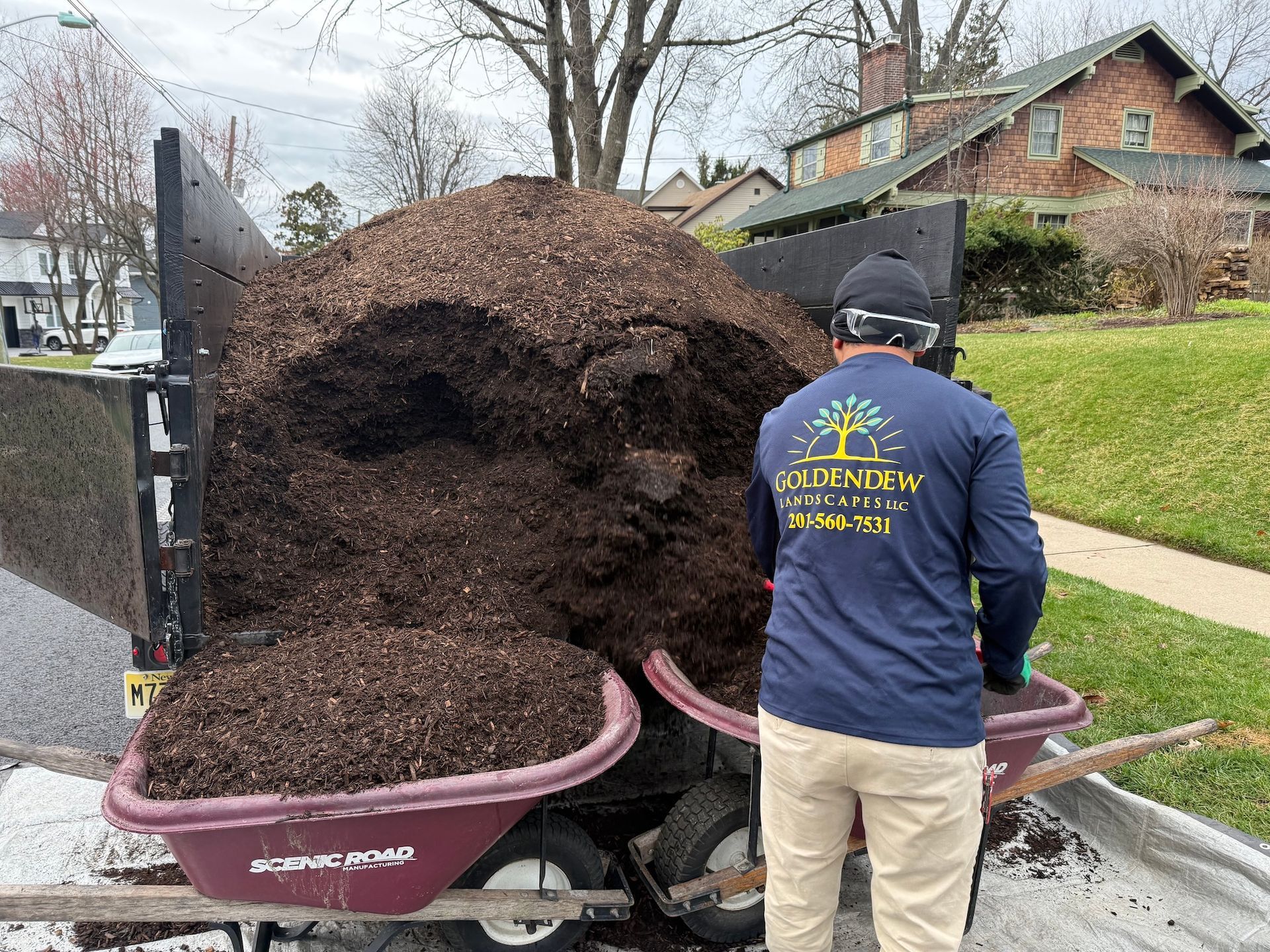 A worker in a navy long-sleeve shirt loads a wheelbarrow with mulch from a trailer parked on a residential street.