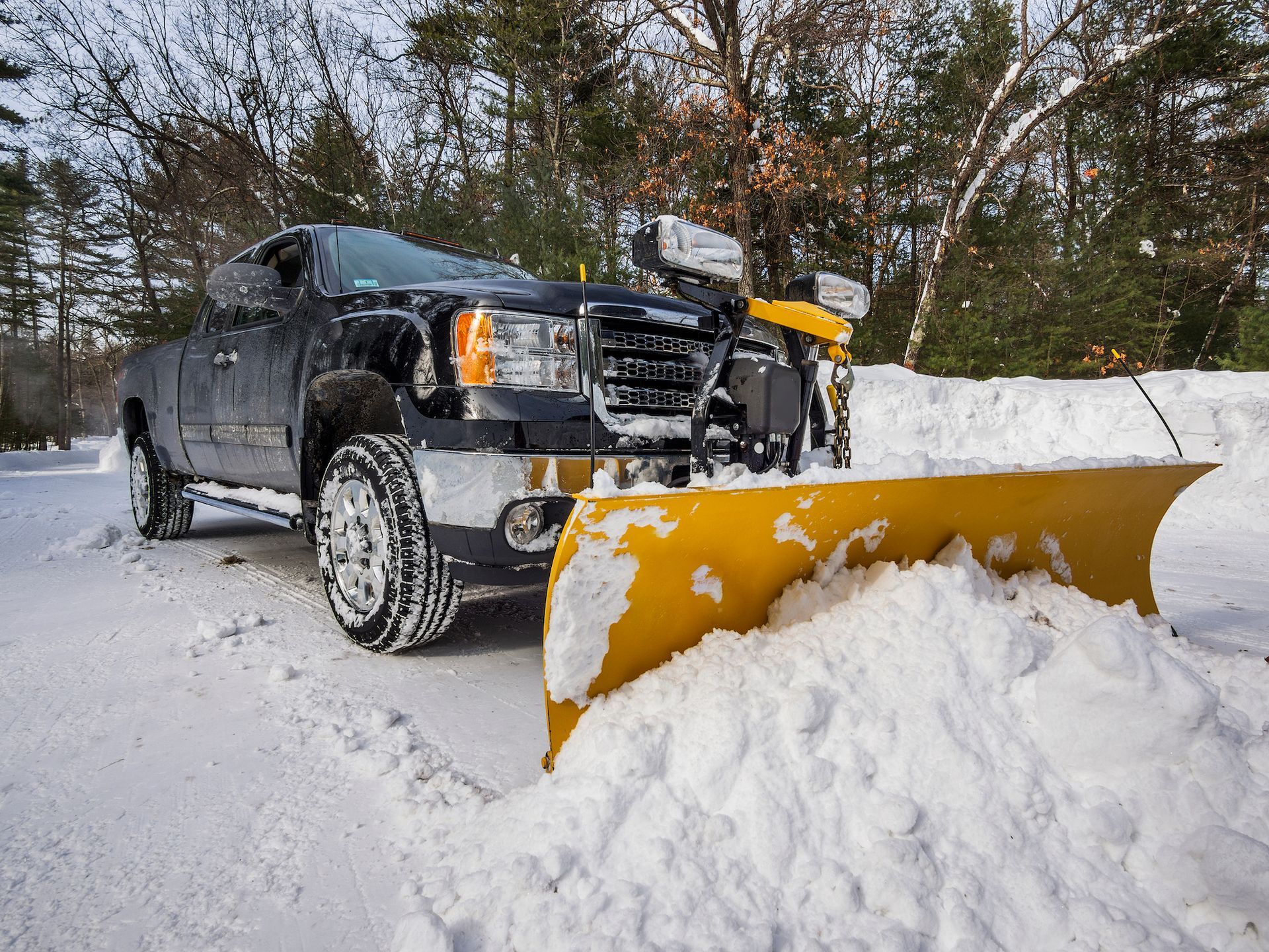 A dark pickup truck with a large yellow snowplow attachment clearing a snowy driveway in a wooded area.