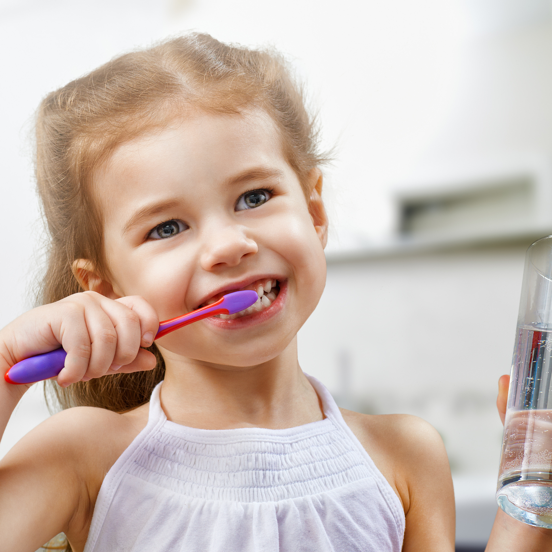 Girl brushing teeth with a pink toothbrush, holding a glass of water, smiling.