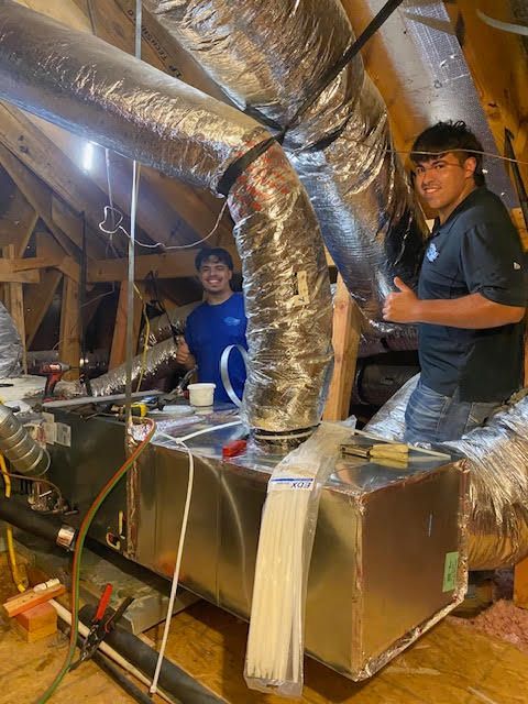 Two men are working on an air conditioner in an attic.