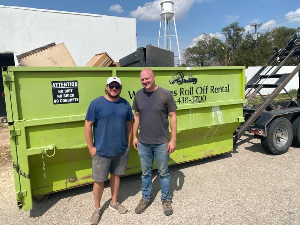 Two men are standing in front of a green dumpster.