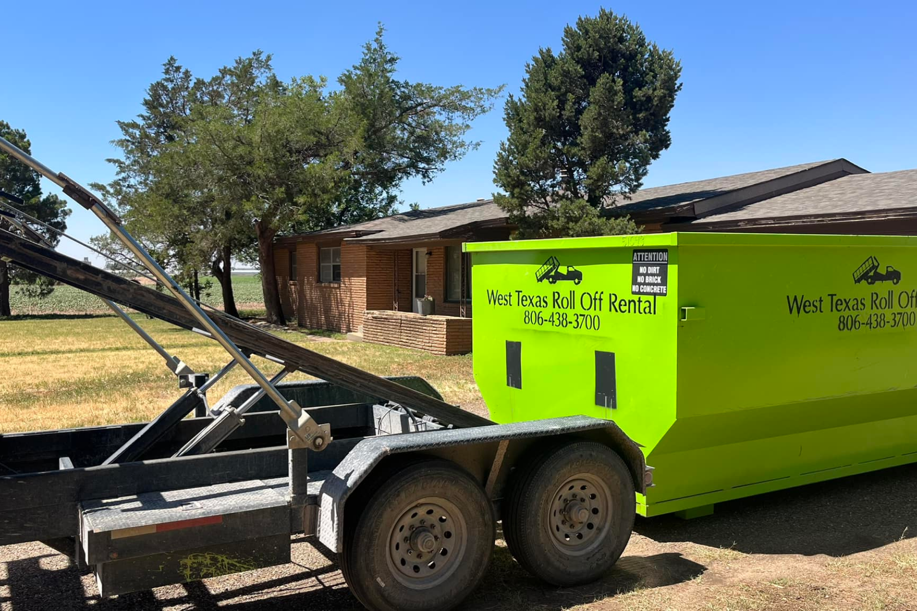 A green dumpster on a trailer is parked in front of a house.