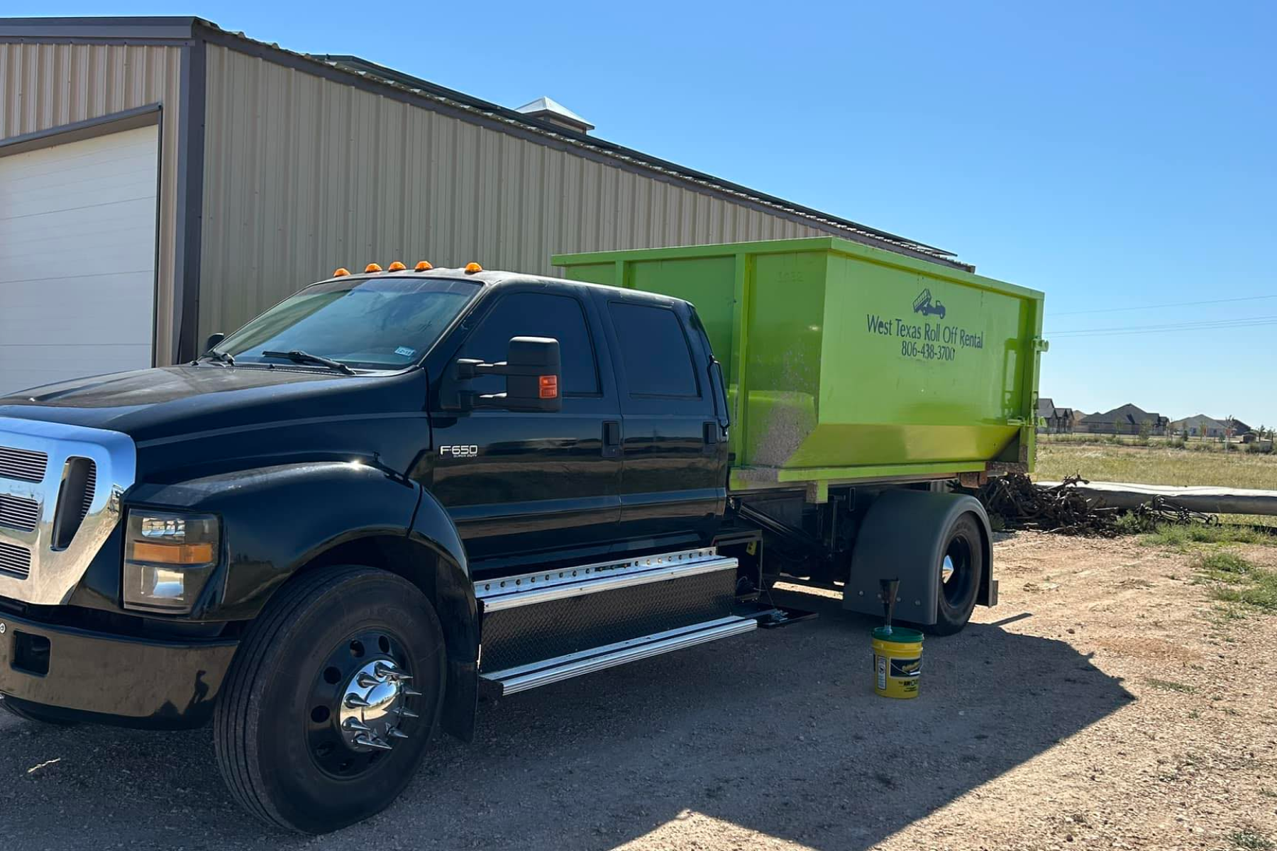 A black truck with a green dumpster attached to it is parked in front of a building.