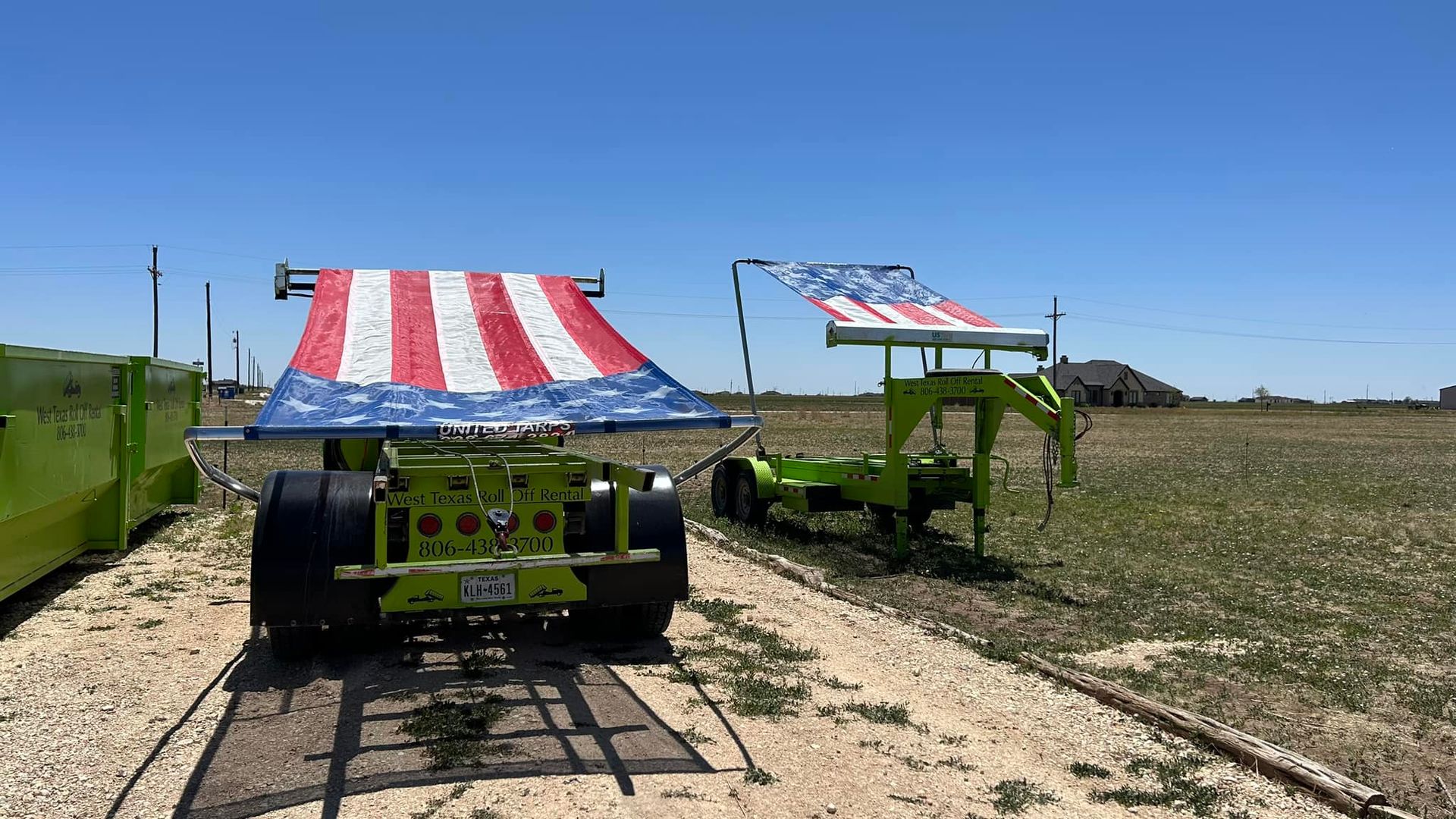 A green truck with an american flag on top of it is parked on a dirt road.