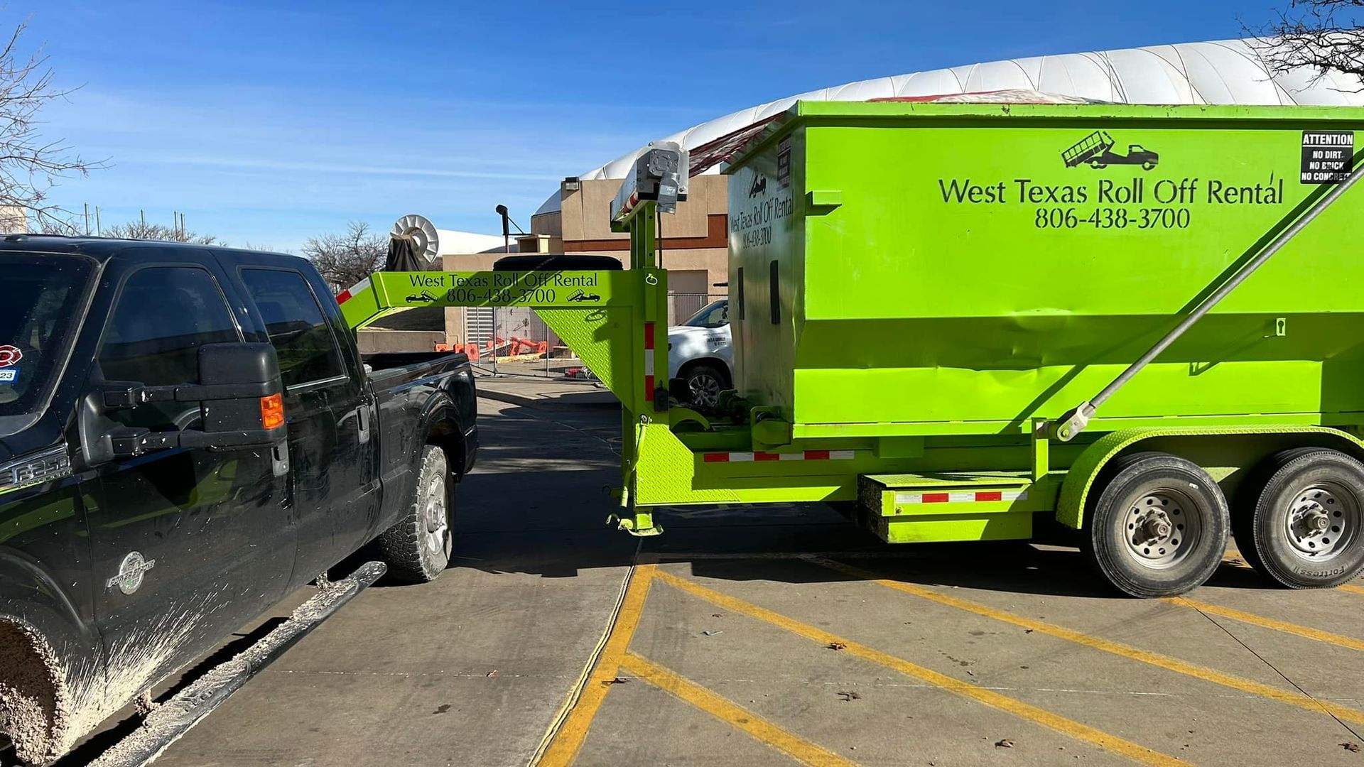 A green dumpster is being towed by a black truck in a parking lot.