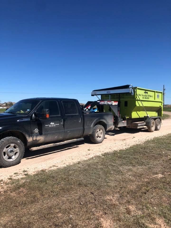 A black truck is towing a green dumpster on a dirt road.