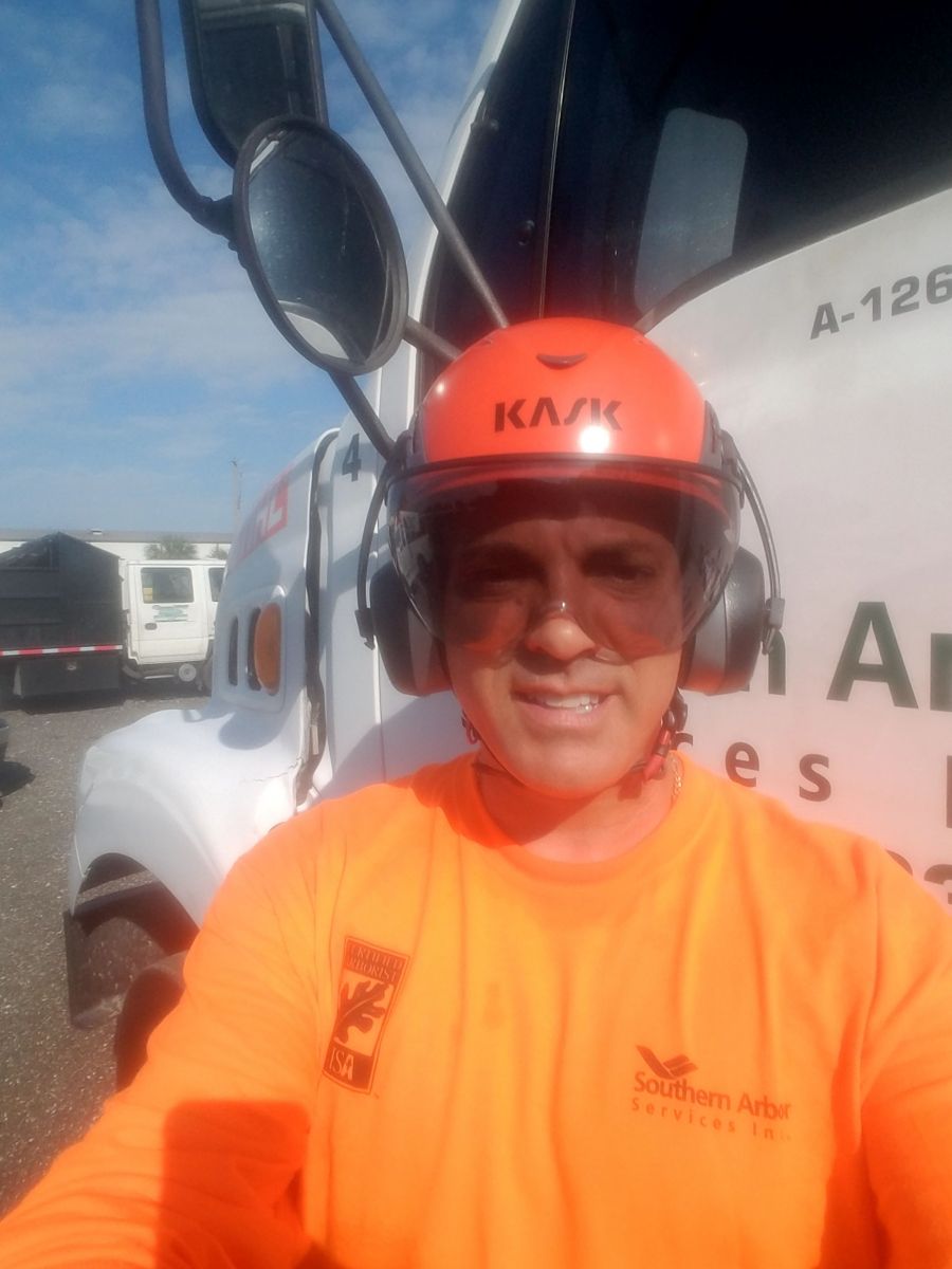 Man in orange helmet and shirt, in front of a truck, wearing ear protection, smiling.