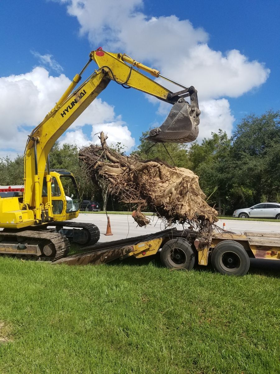 Yellow excavator loads a large pile of dirt and debris onto a flatbed trailer on a grassy area, under a blue sky.
