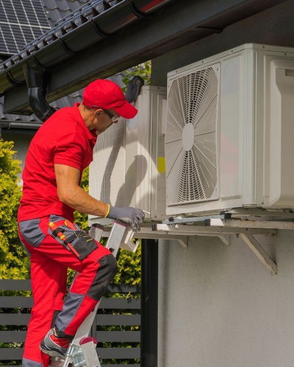 A man is standing on a ladder fixing an air conditioner on the side of a building.