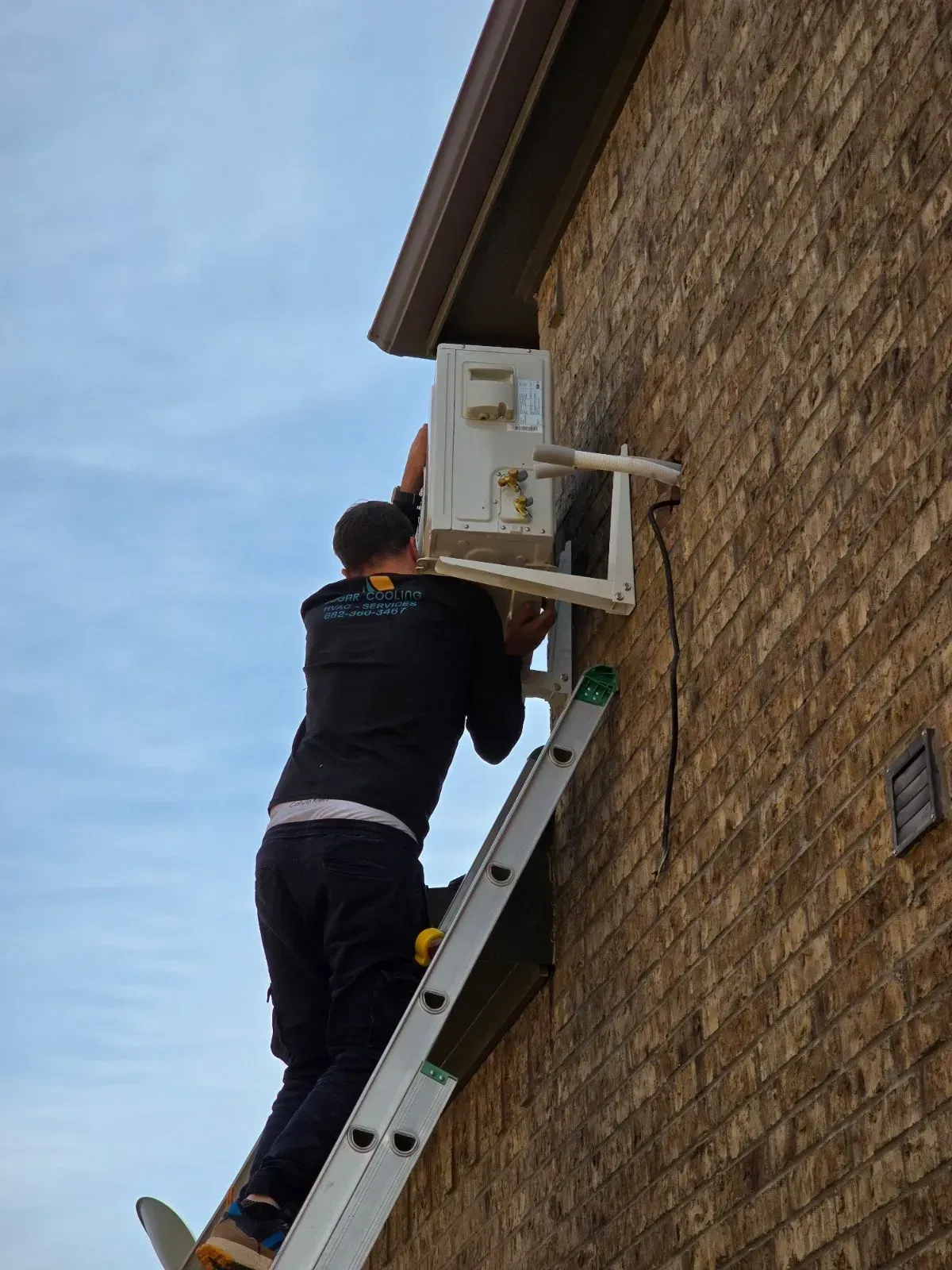 A man is climbing up a ladder to install an air conditioner on the side of a brick building.