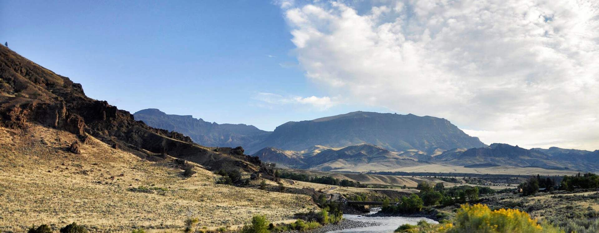 A river flows through a dry, grassy valley leading toward rugged, dark mountains under a bright blue, cloudy sky.