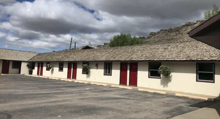 Single-story motel with a weathered wood-shake roof, light-colored walls, red doors, and a parking lot under a cloudy sky.