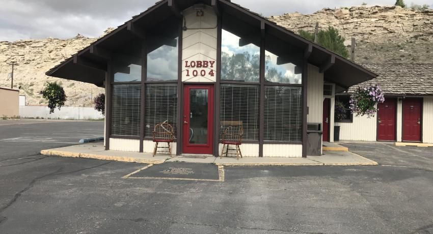 A motel lobby with a red front door, large windows, and an overhang, set against a rocky hill under a cloudy sky.