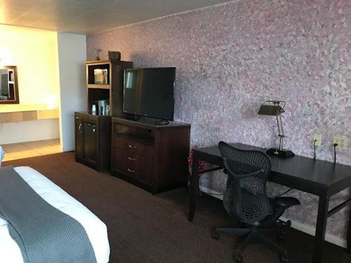 A hotel room with a bed, a television on a wooden dresser, and a desk with a black mesh office chair against a textured wall.
