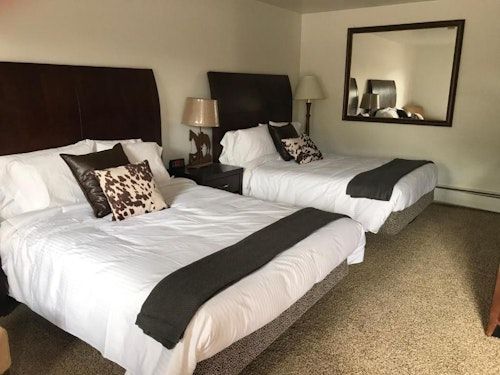 A hotel room containing two queen beds with dark headboards, white linens, and patterned accent pillows on beige carpet.