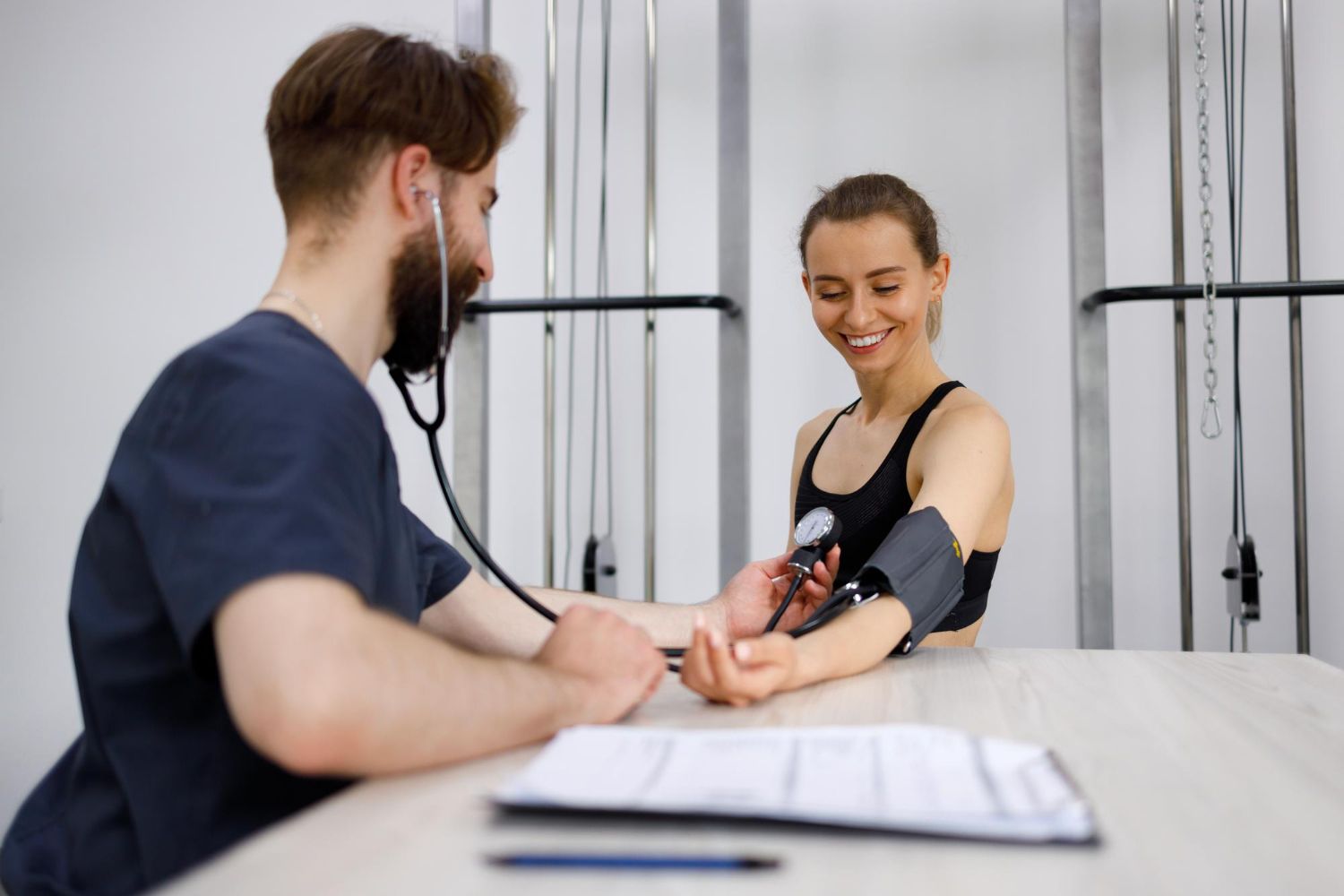 A healthcare professional uses a stethoscope and cuff to measure the blood pressure of a smiling person at a table.