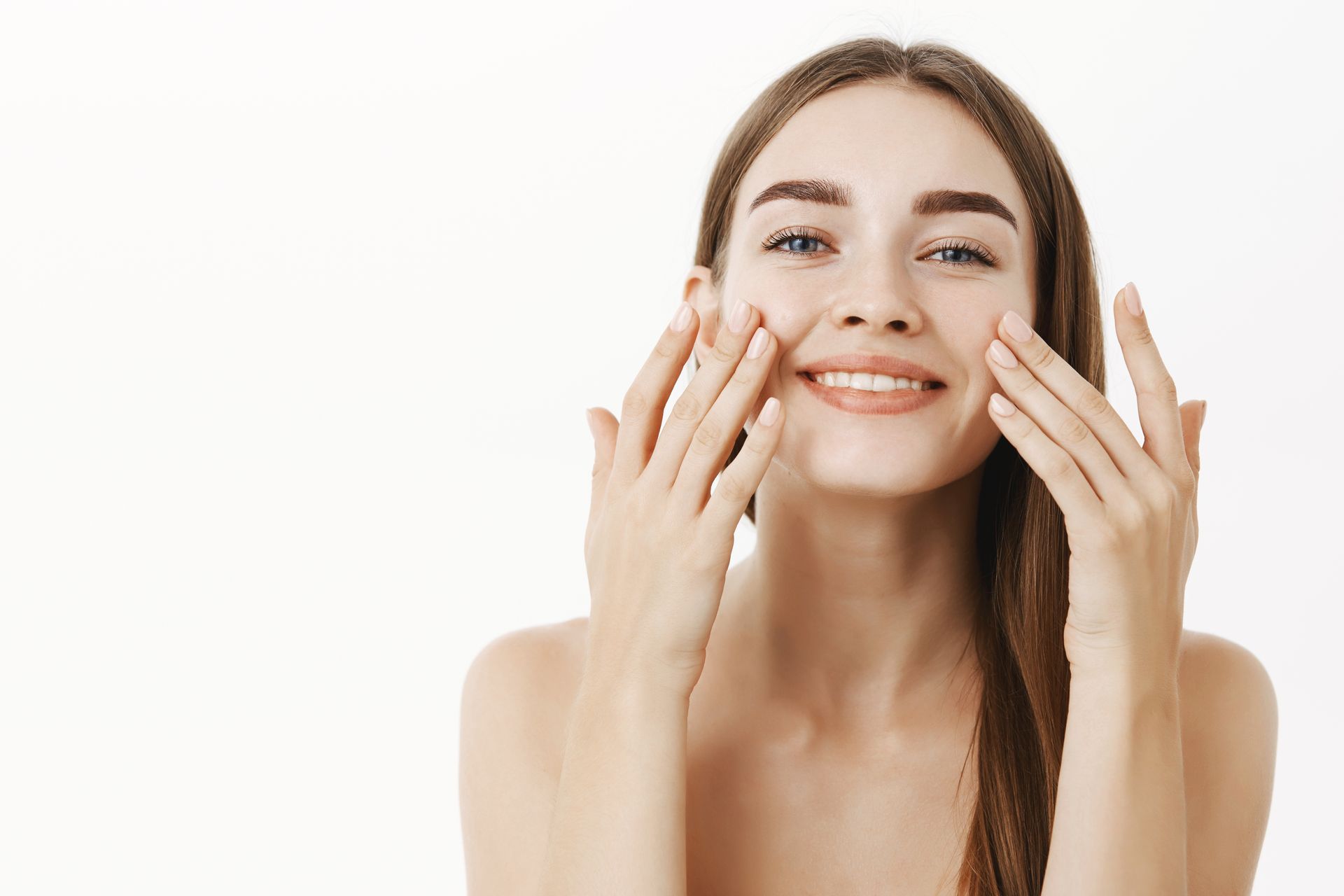 A person with smooth skin smiling while gently touching their cheeks against a white background.