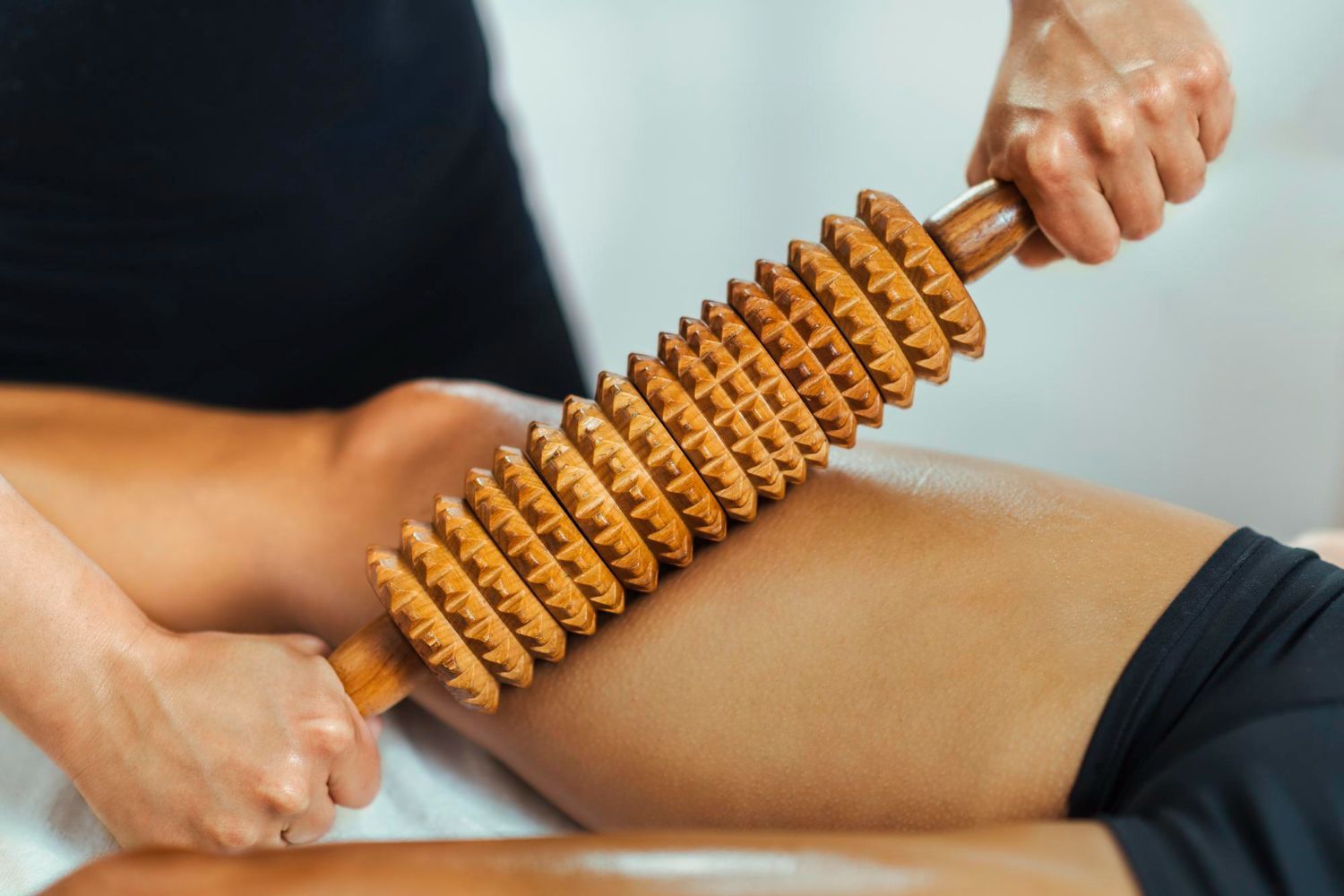 A wooden massage roller with textured wheels is being used to massage a person's thigh.