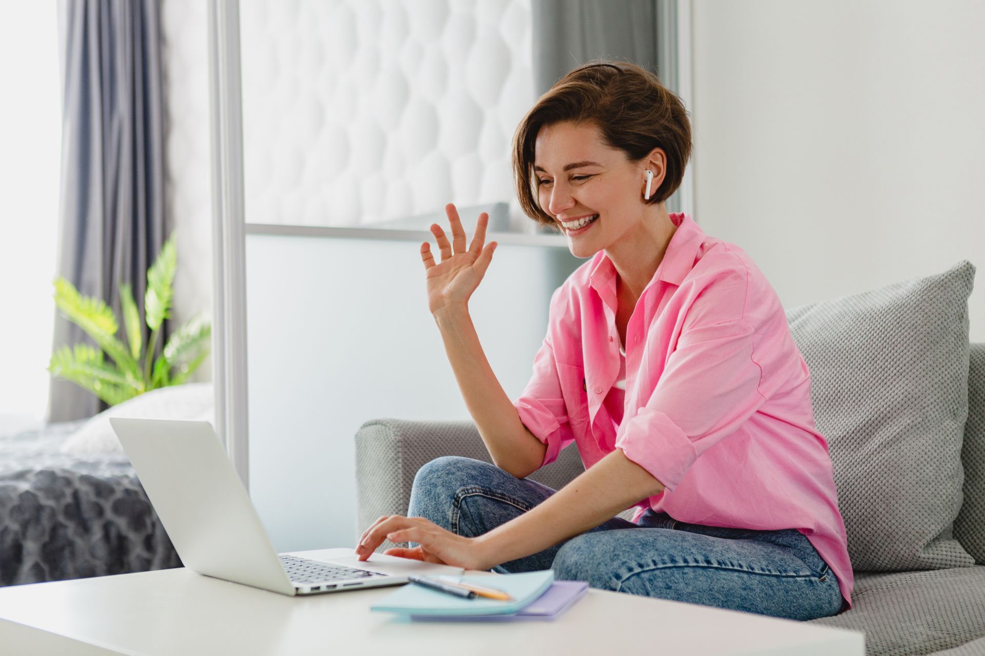 Woman seated on a couch, using laptop, waving, and smiling; pink shirt, blue jeans, white earbuds.