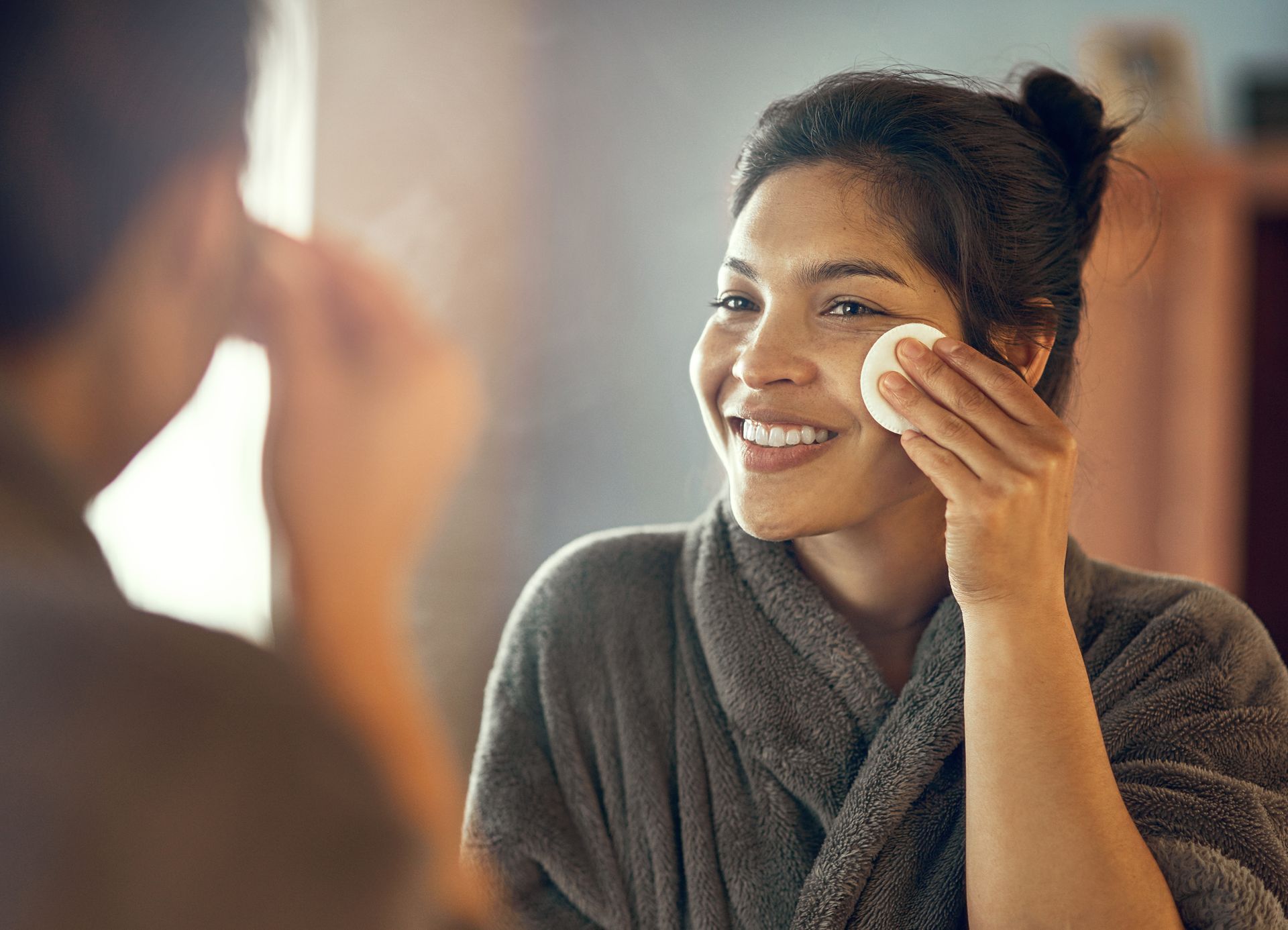 A woman is smiling while cleaning her face in front of a mirror.