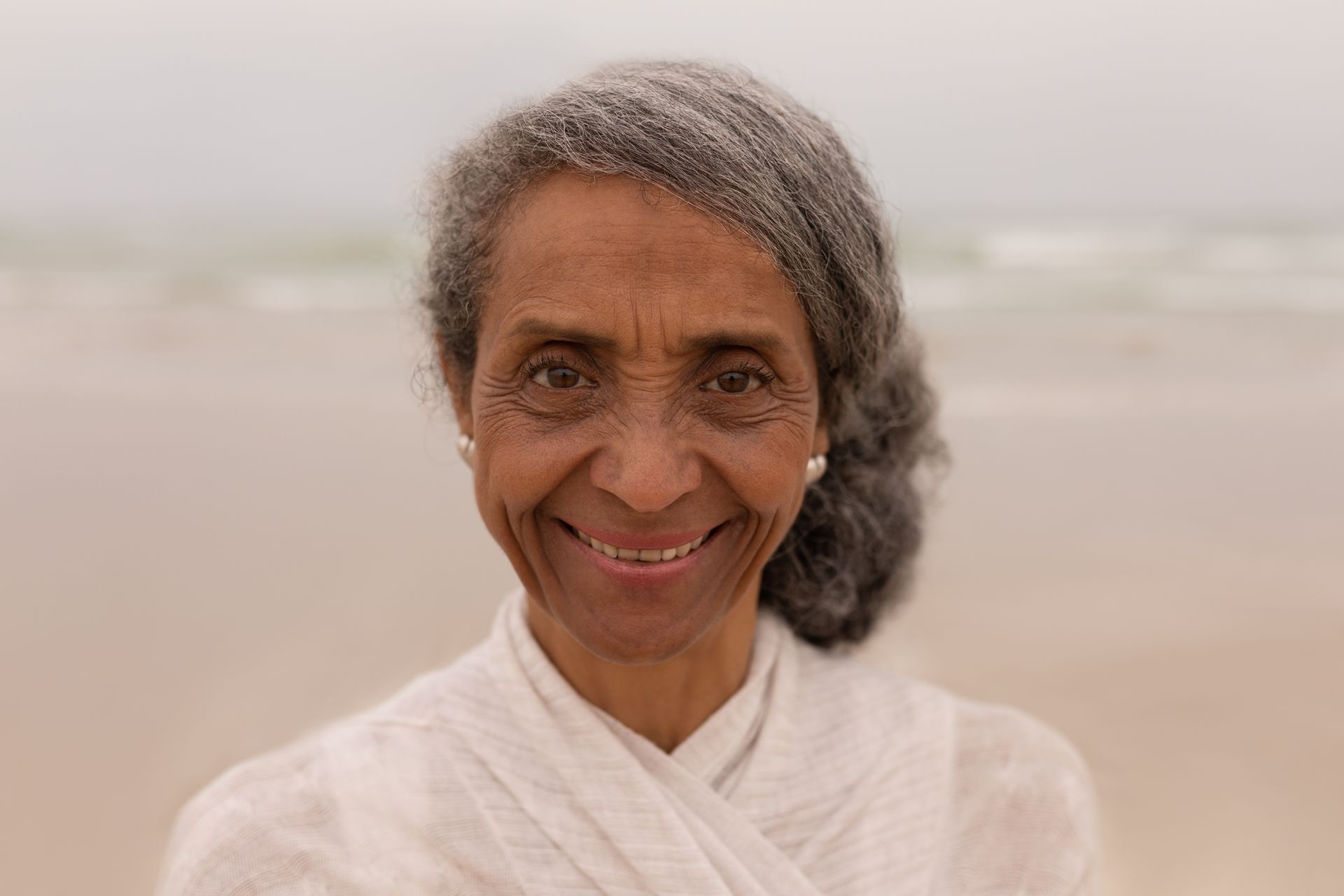 Smiling elderly woman with gray hair at the beach, wearing white clothing.