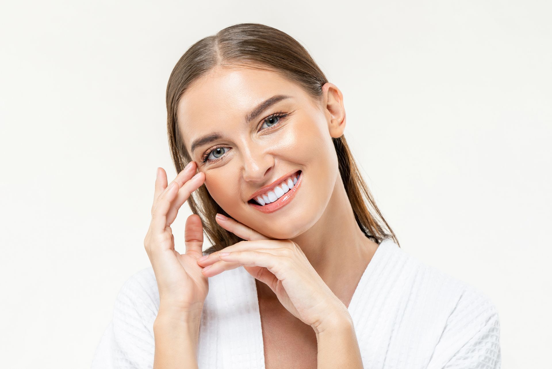 Woman in white robe smiles, touching her face, with bright skin and a light background.