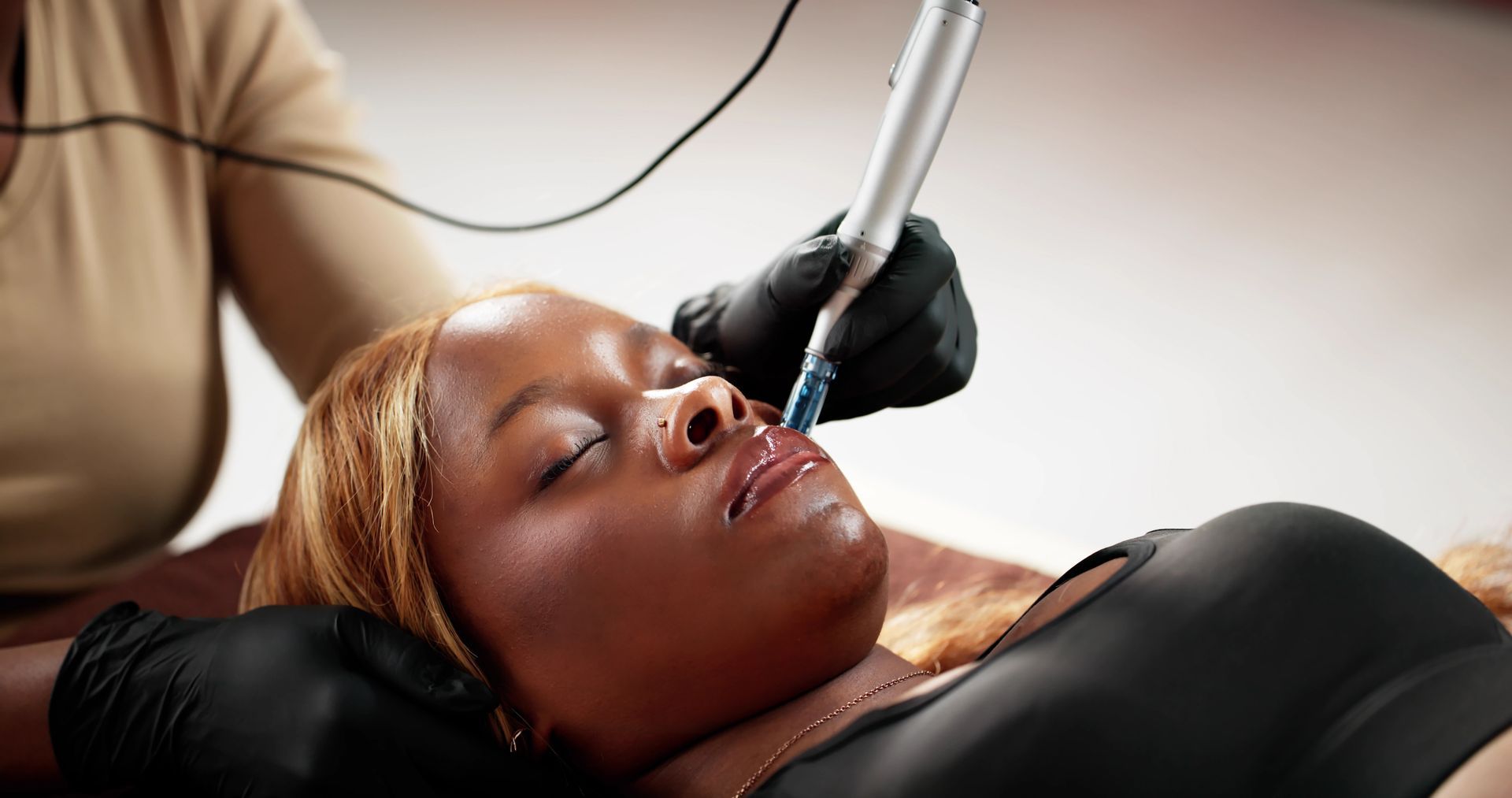 Woman receiving lip tattoo, lying down. Black gloved hand uses a tattoo pen on her lip.