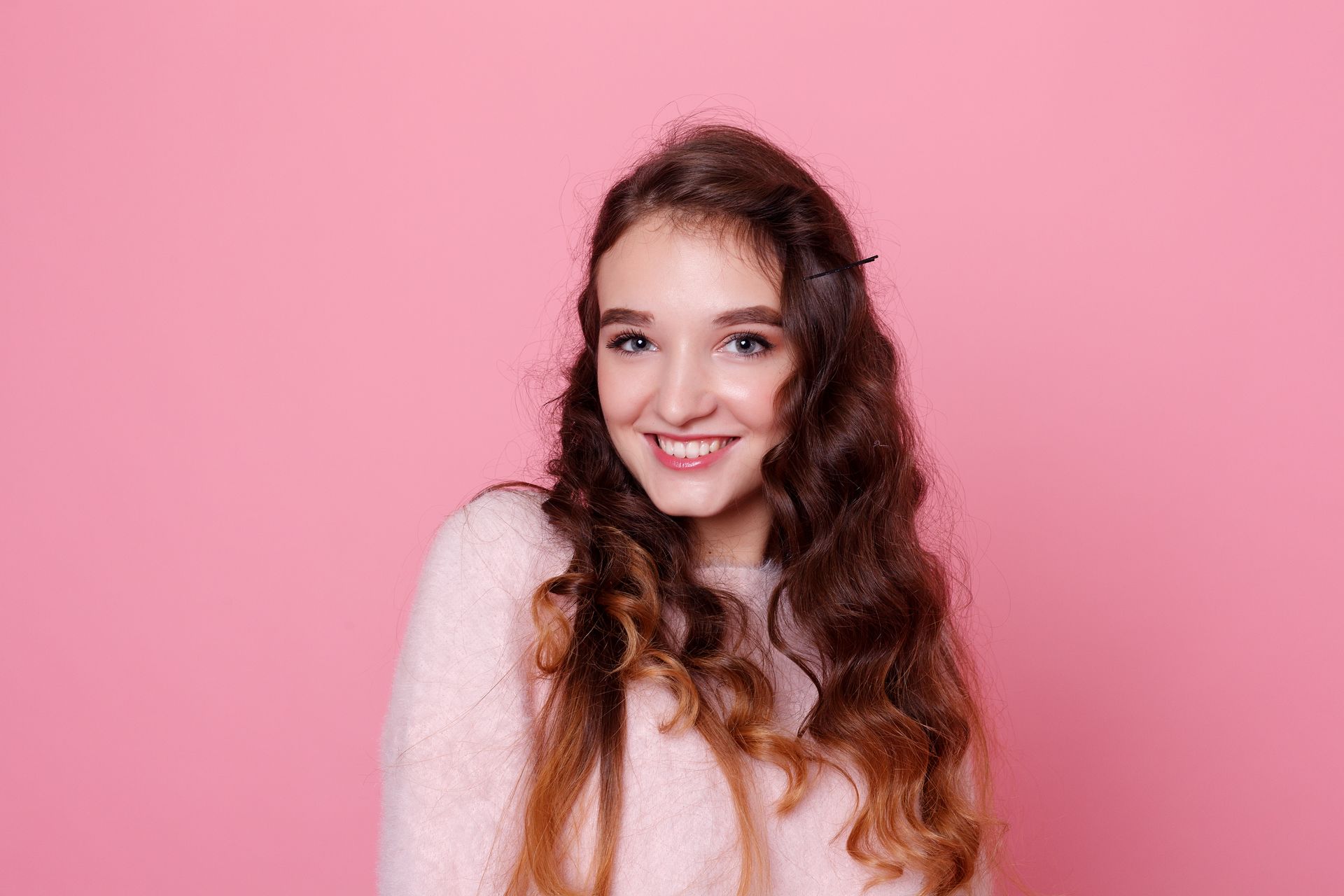 Woman with long, wavy brown hair smiles at the camera, wearing a pink sweater, in front of a pink background.