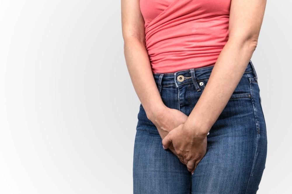 Woman in jeans and pink top holding her hands near her lower abdomen, likely indicating discomfort.