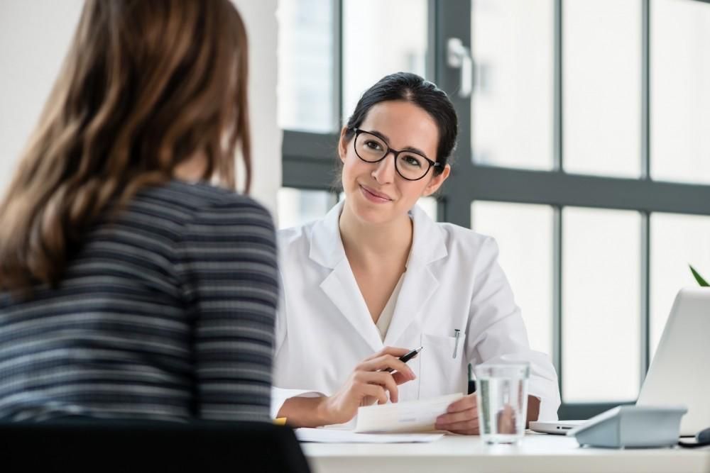 Doctor in white coat consults with a patient; they are seated at a desk, looking at paperwork.
