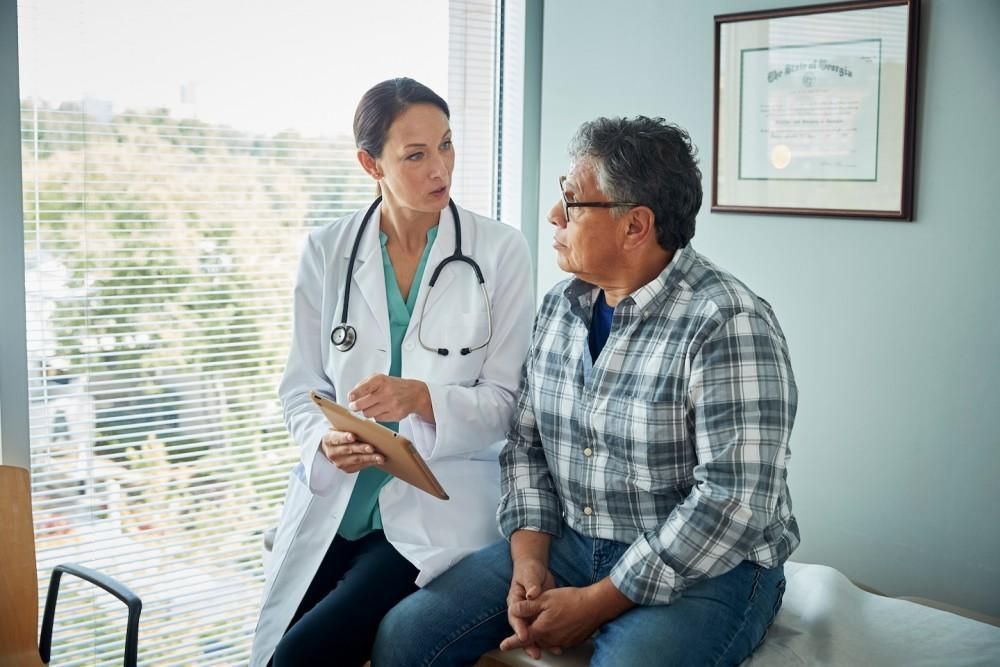 Doctor in white coat speaking with patient, holding a clipboard in an exam room.