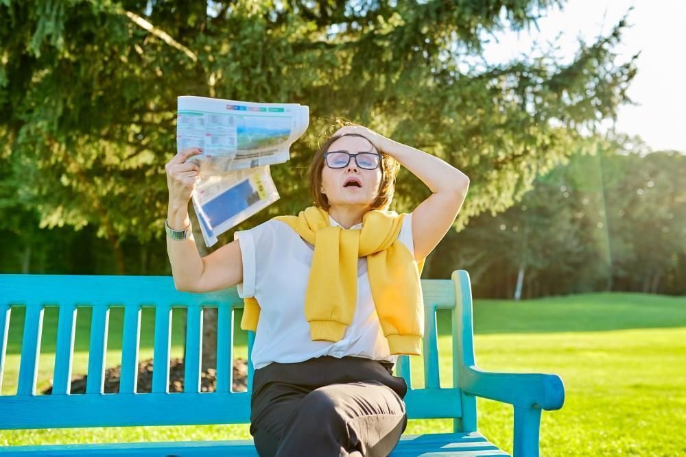 Woman on blue bench fanning herself with newspaper in sunny park.