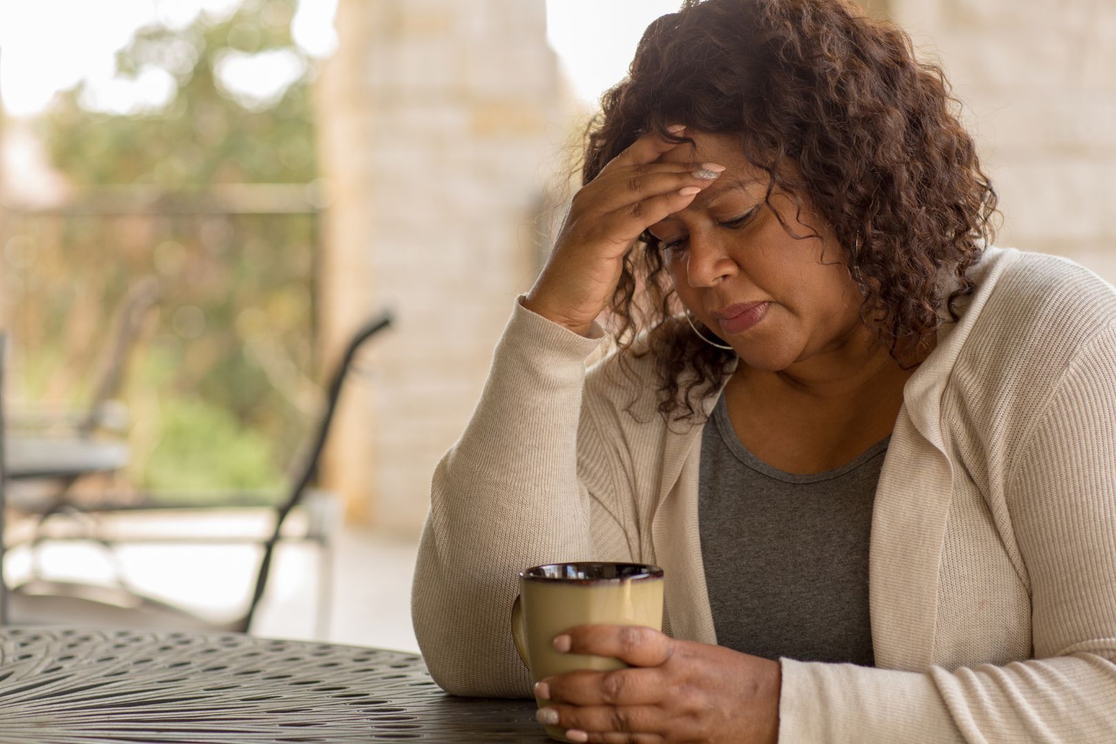 Woman holding a mug, resting her hand on her forehead, looking down with a pensive expression, seated outdoors.