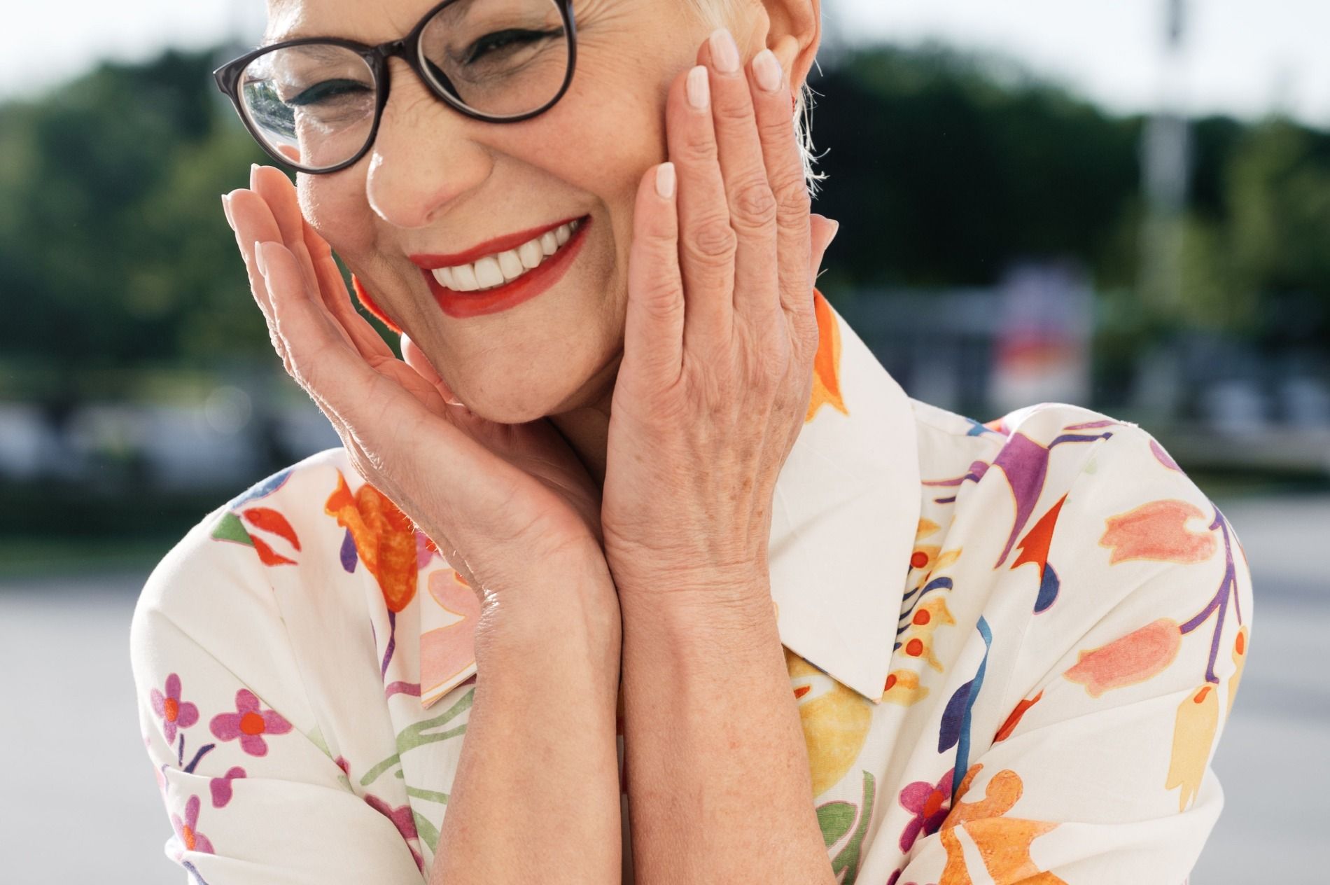 Woman with glasses smiles, hands on her cheeks; floral shirt, outdoors.