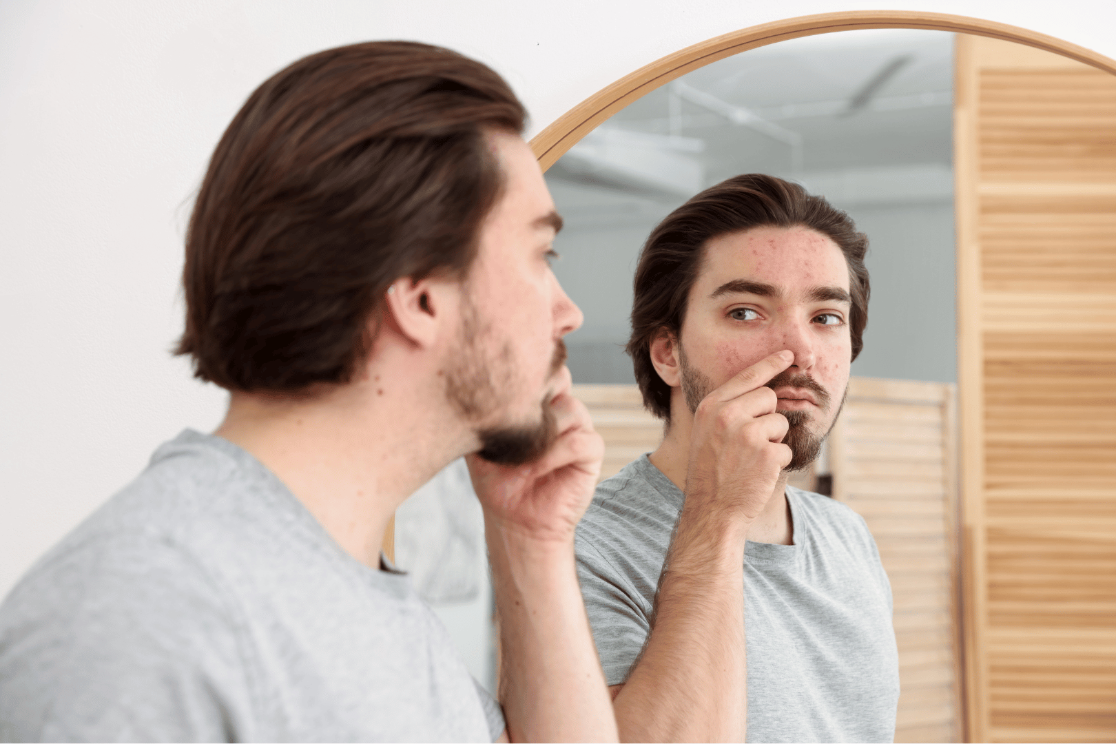 Man looking in mirror, touching his nose; red spots visible.