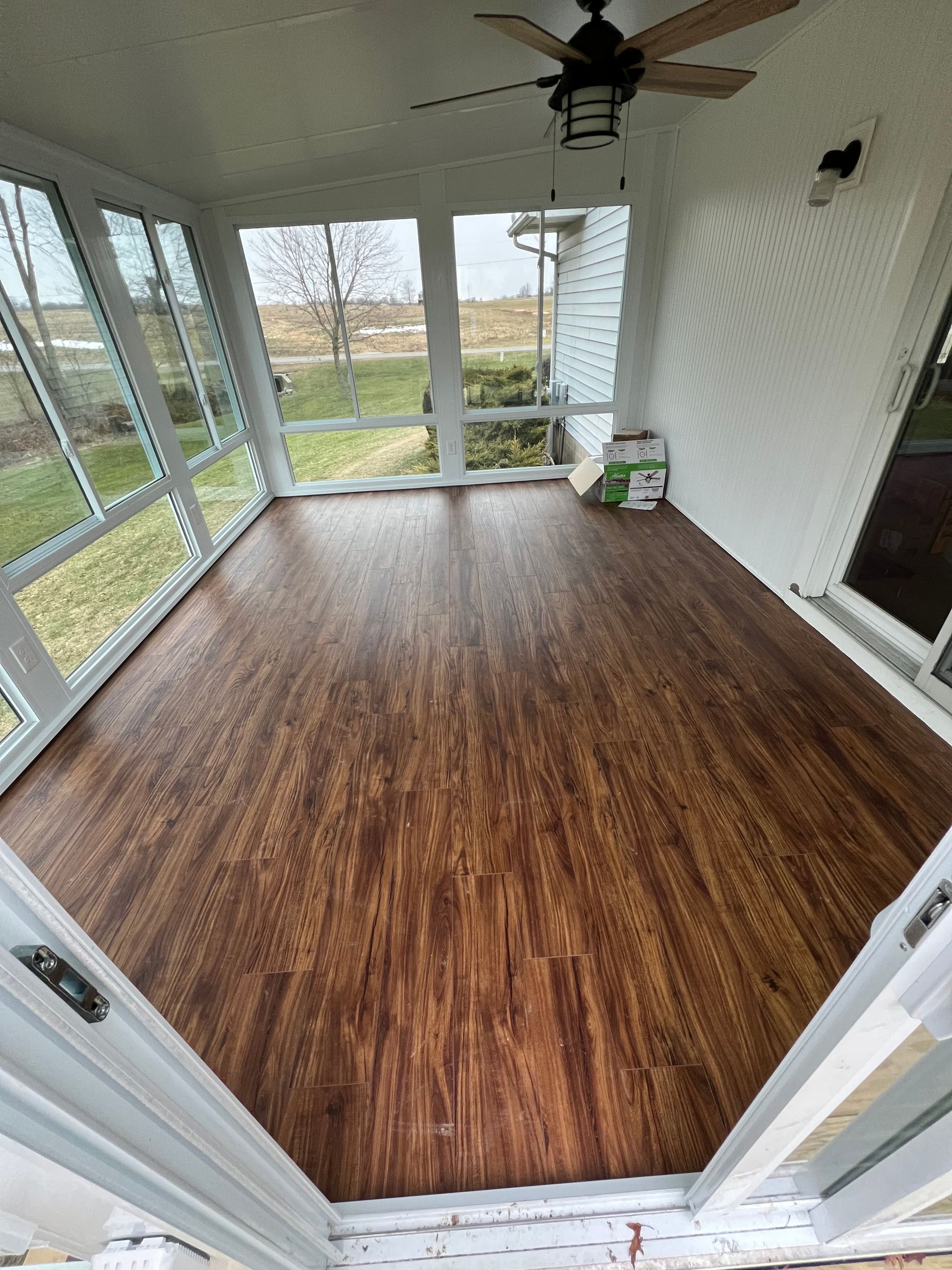 A sunroom featuring dark wood-look plank flooring, large windows, a ceiling fan, and white walls.