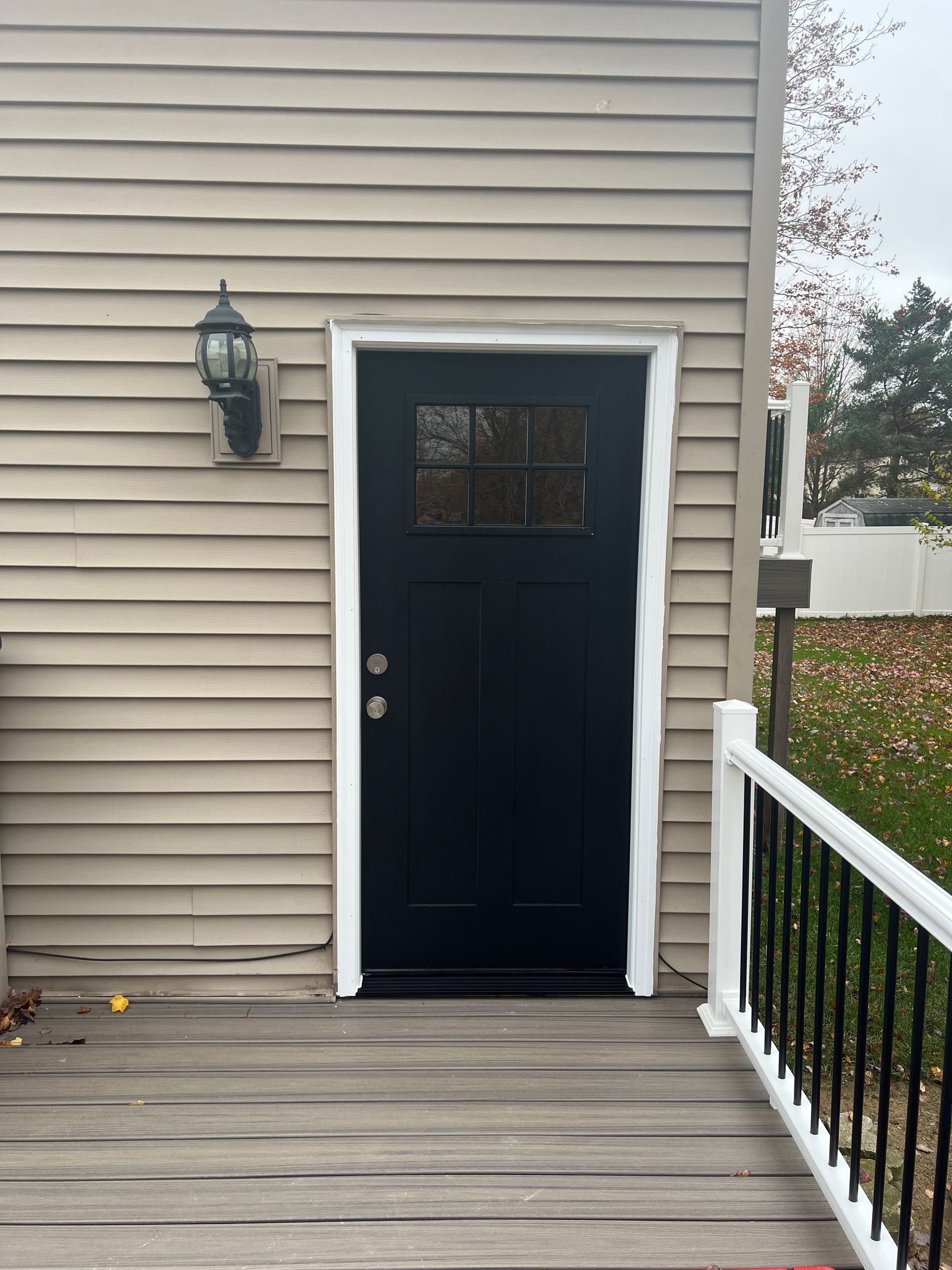 A black door with a glass pane window in a tan siding wall, featuring a wall-mounted light and a white deck railing.