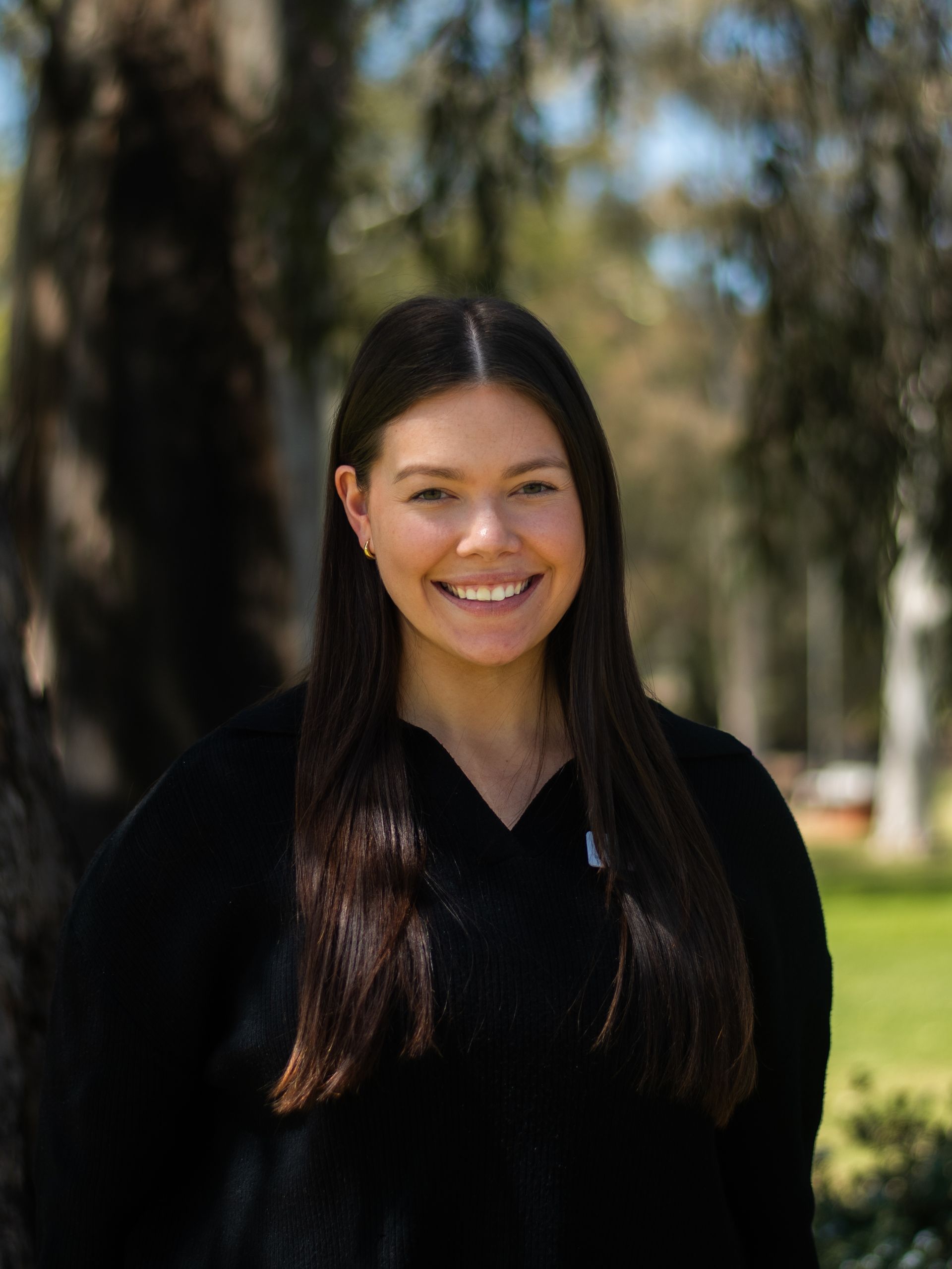 A woman in a black sweater is smiling in front of a tree.
