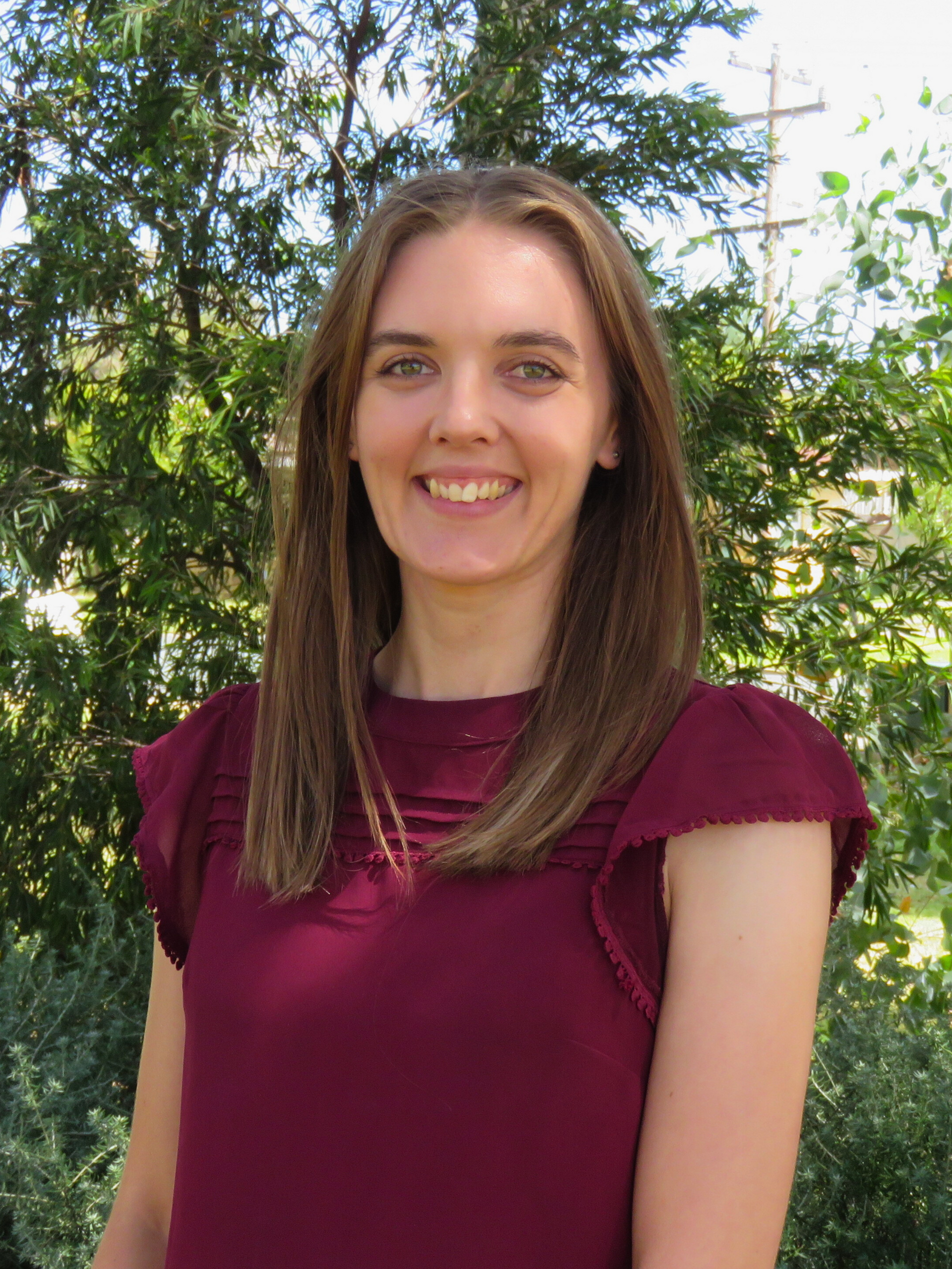 A woman in a red shirt is smiling for the camera while standing in front of trees.
