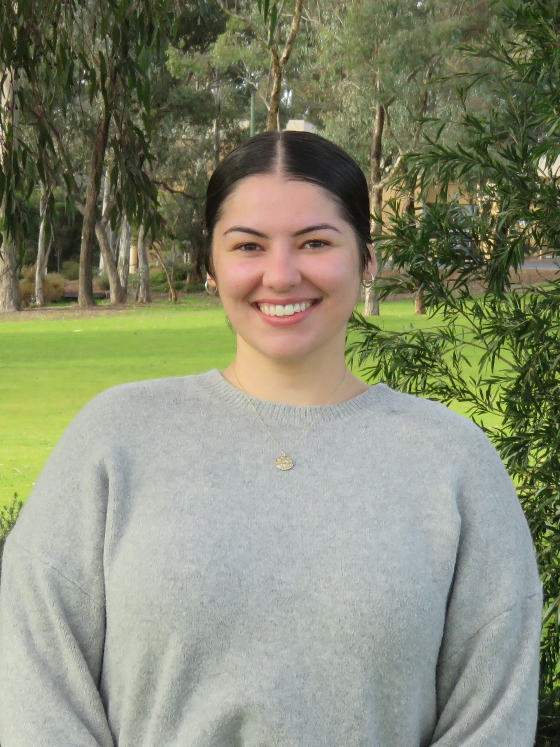 A woman in a grey sweater is smiling in front of trees.