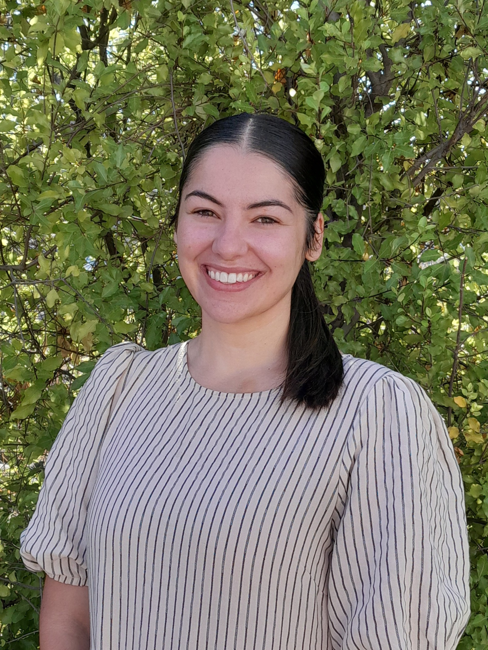 A woman in a grey sweater is smiling in front of trees.