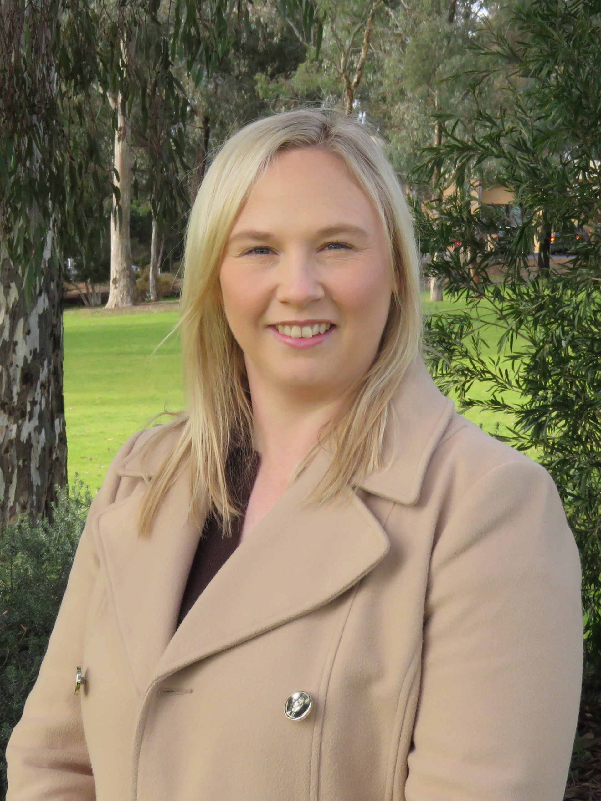 A woman in a tan coat is smiling in front of a tree.