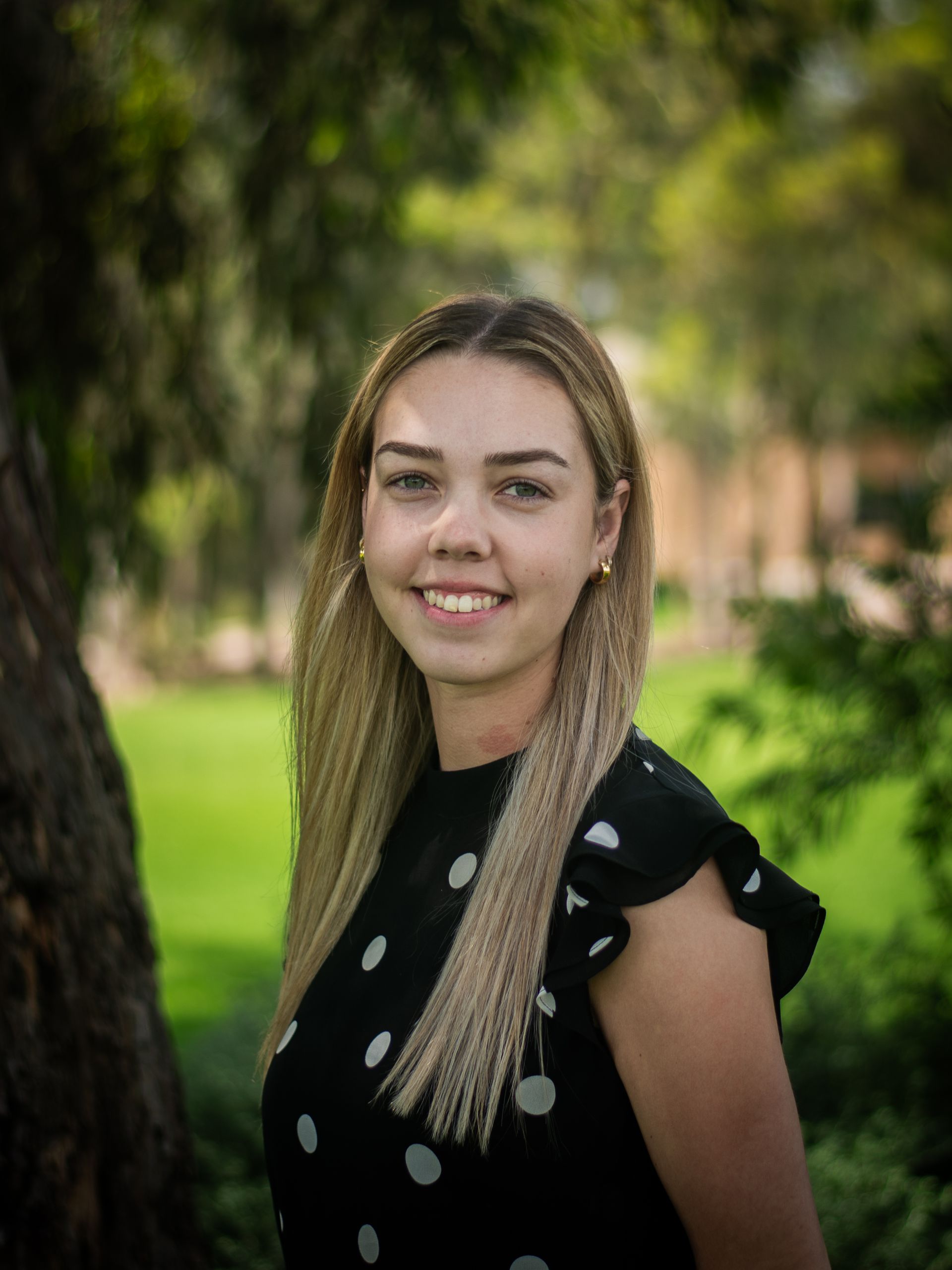 A woman in a black polka dot dress is standing next to a tree.