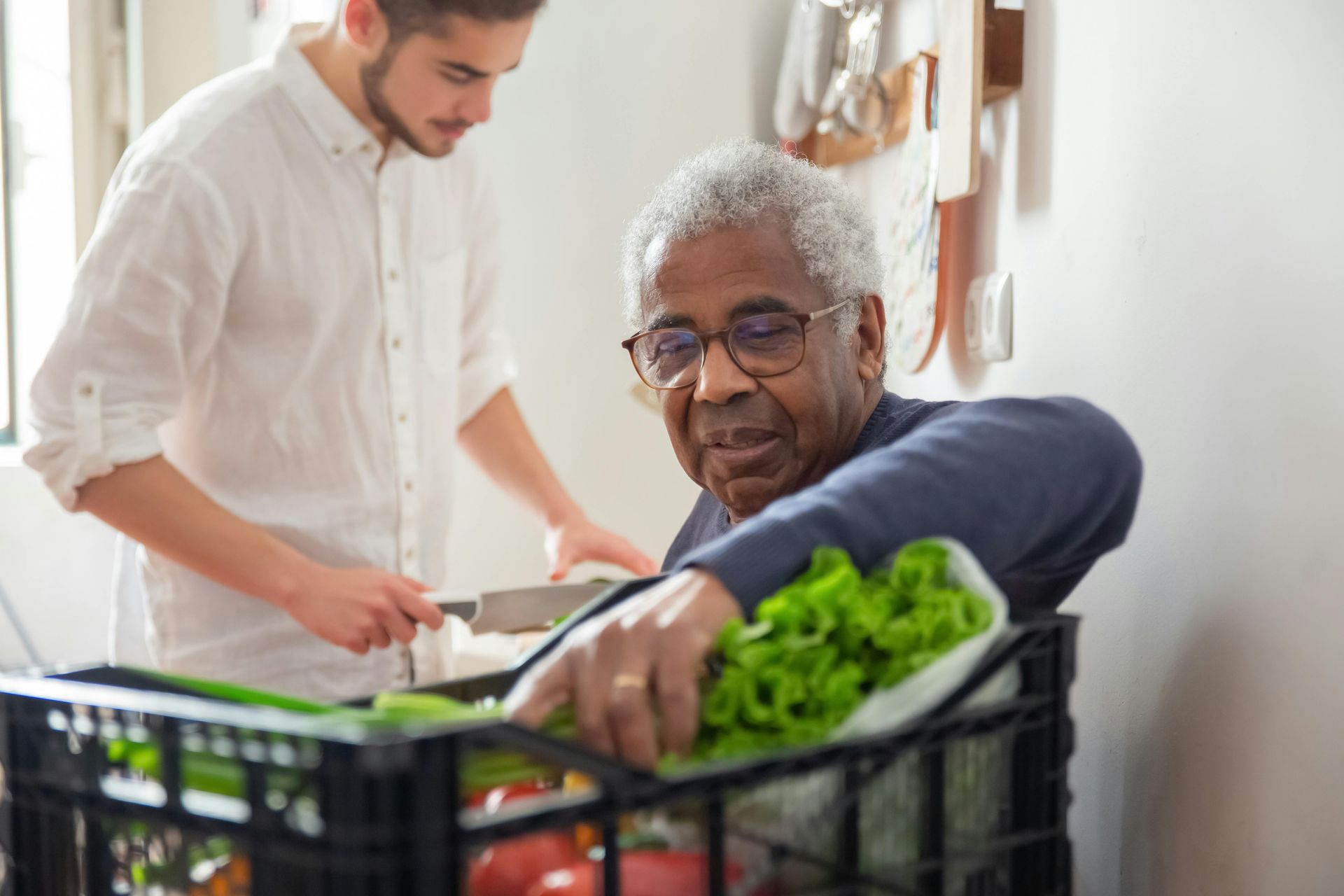 Man in glasses reaches for vegetables in a basket while another person prepares food nearby.