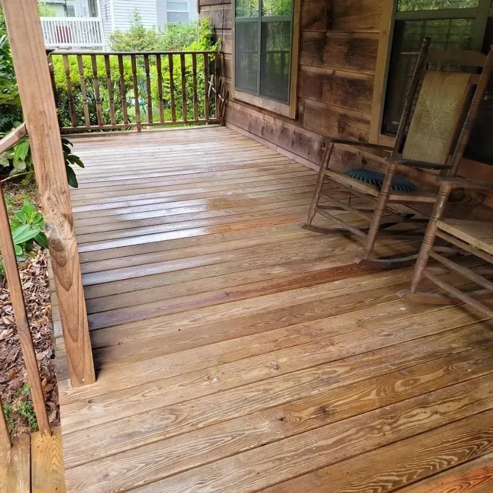 Wooden porch with two rocking chairs, railing, and a window. Sunlight shines on the weathered wood.
