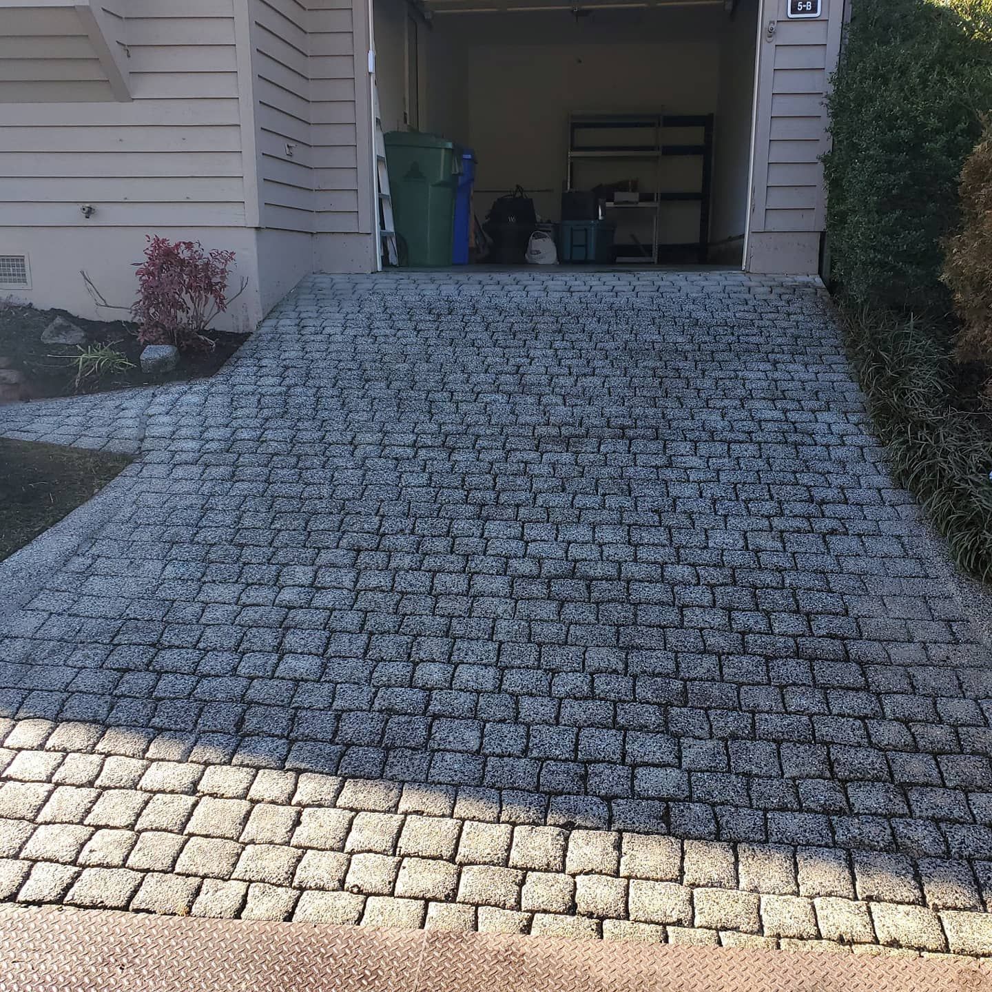 Cobblestone driveway leading up to a garage. A green trash can and various items are visible in the garage.