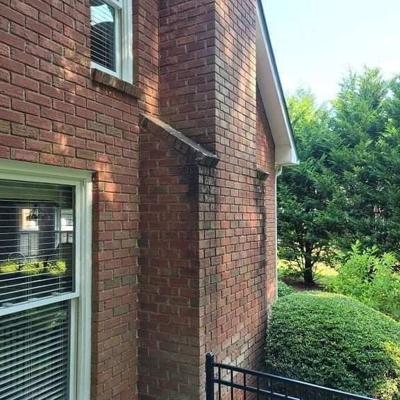 Brick chimney and exterior wall with dark streaks, next to greenery and a window.