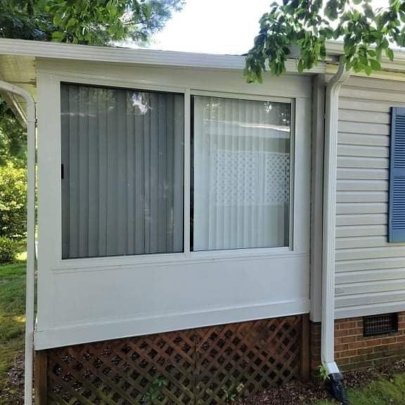 White sunroom with vertical blinds, attached to a gray-sided building with blue shutters.