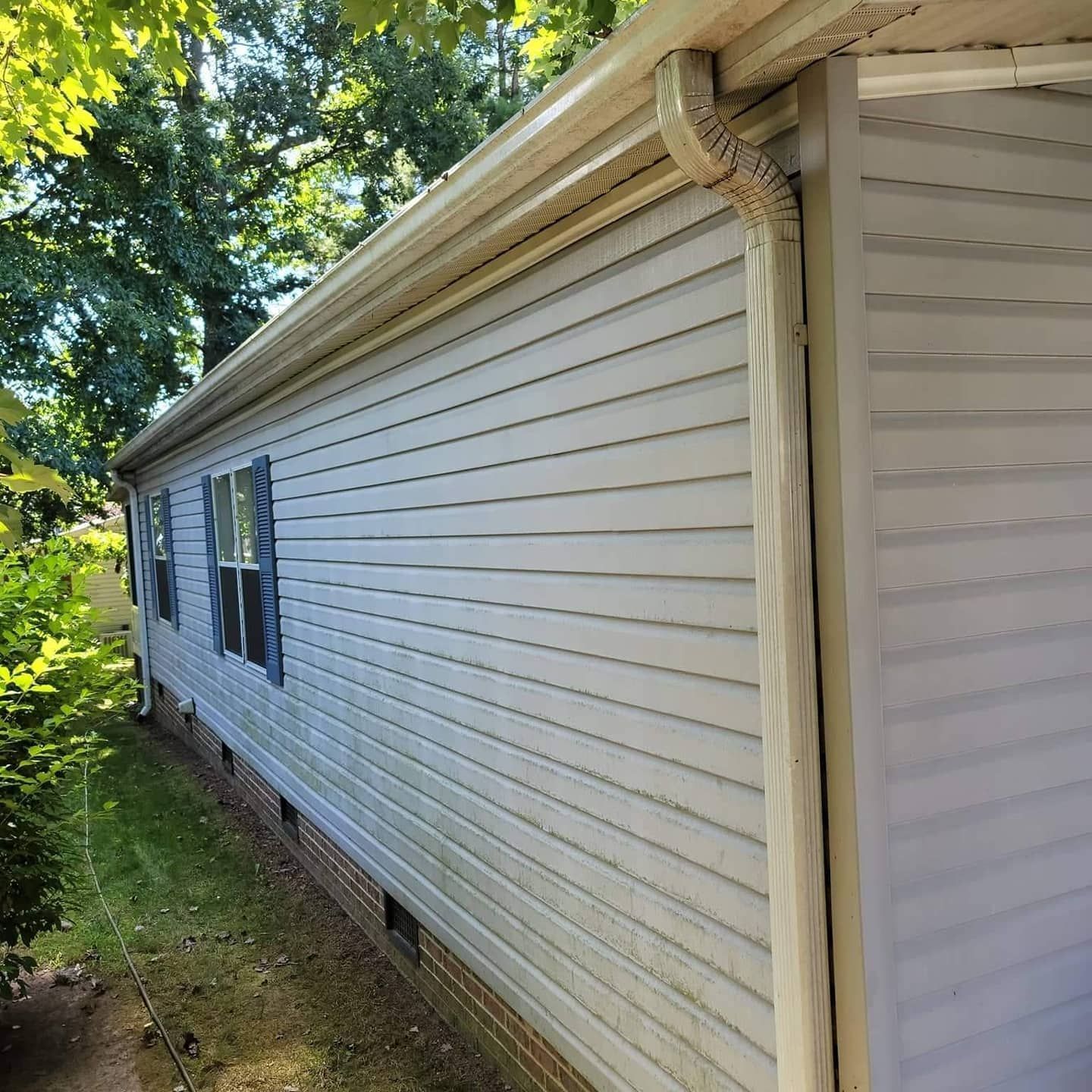 Beige siding on a house with gutters and a downspout, surrounded by green foliage.